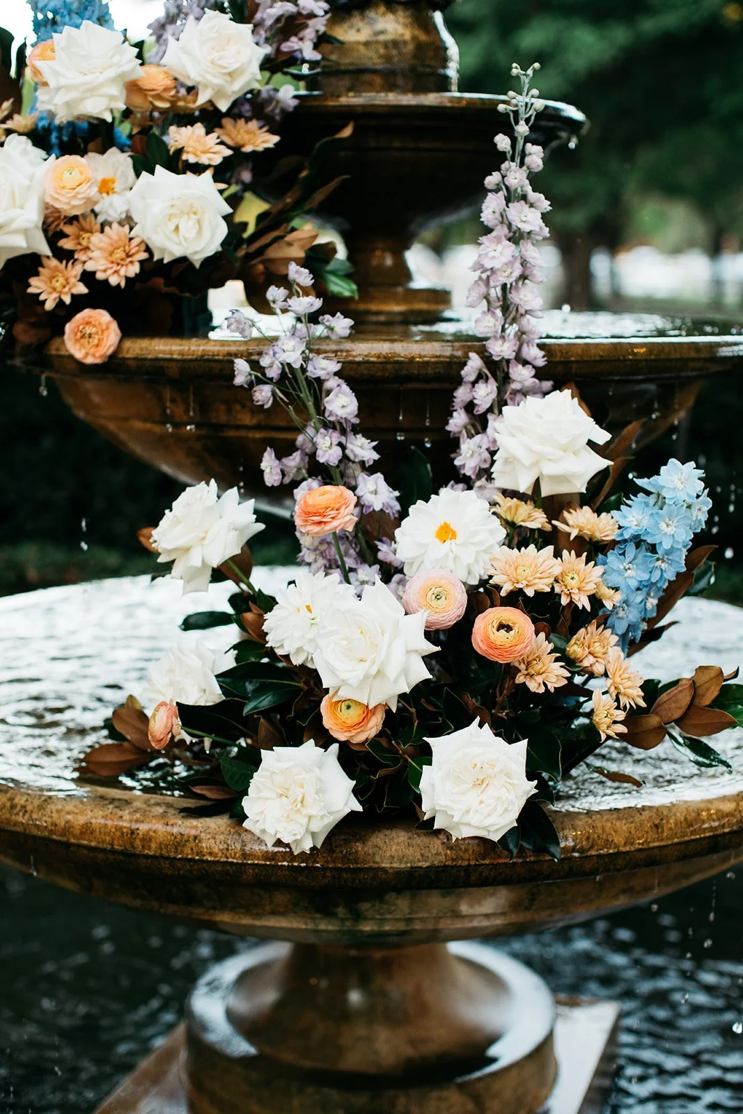A decorative water fountain adorned with an arrangement of various pastel-colored flowers including white, peach, light purple, and blue, with greenery.