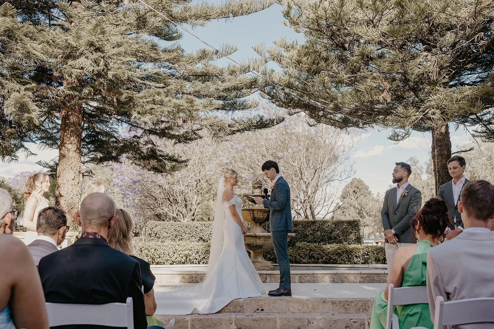 A wedding ceremony taking place outdoors under large trees with a water fountain in the background. The bride and groom stand in front of an officiant, with bridesmaids and groomsmen nearby. Guests are seated and watching the ceremony.