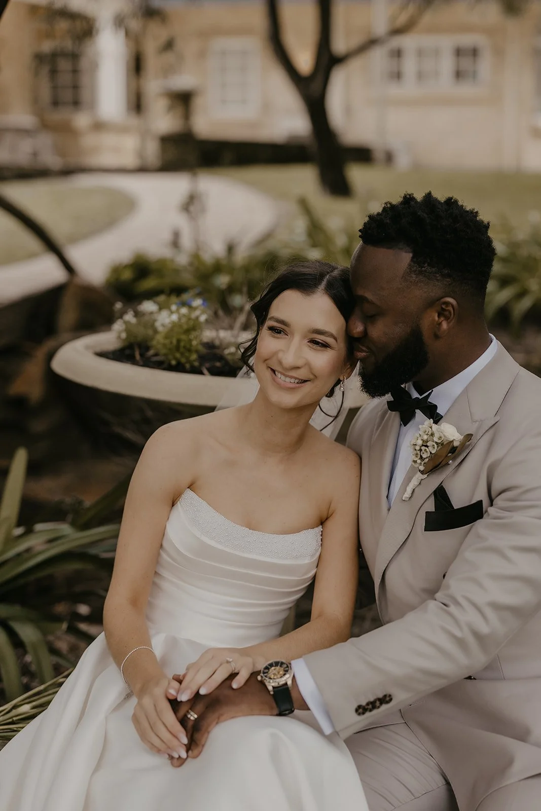Smile wedding couple sitting outdoors, woman in wedding dress, man in beige tuxedo with black bow tie, holding hands, romantic moment.
