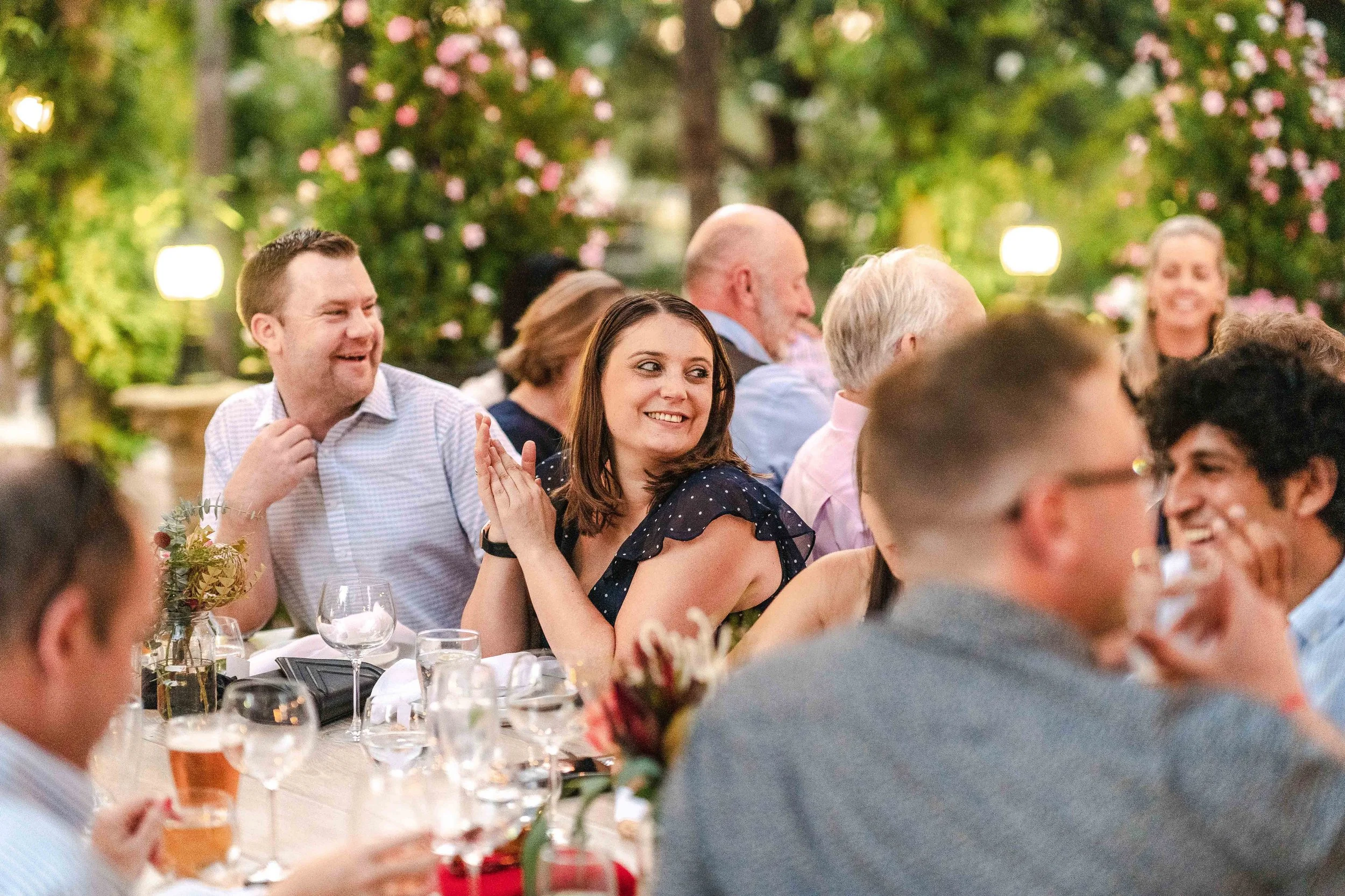 people sitting at a banquet table outdoors, smiling and enjoying a social gathering in a garden setting with greenery and flowering trees in the background.