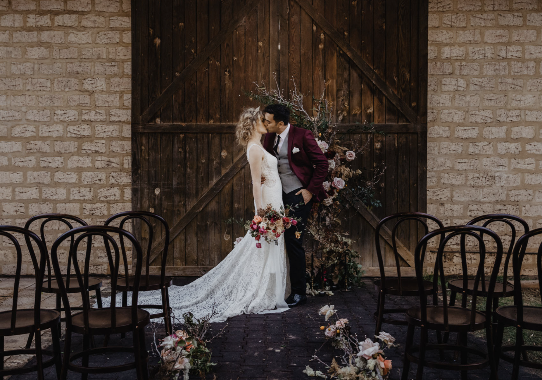 A bride and groom share a kiss in front of a rustic wooden barn door decorated with flowers and greenery, with empty chairs arranged for a wedding ceremony in the foreground.