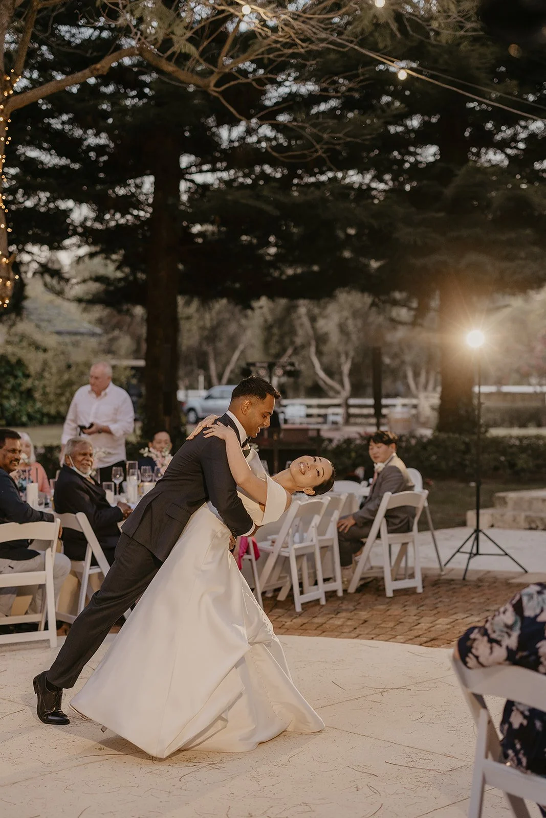 A bride and groom sharing a dance at their wedding reception outdoors during sunset, with guests sitting at tables in the background.