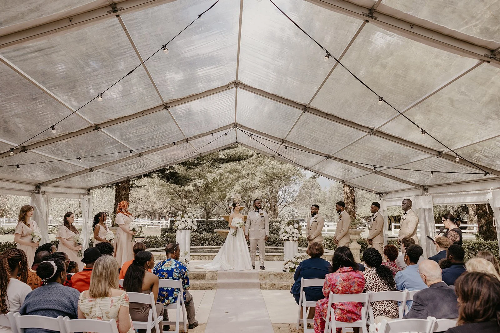 Outdoor wedding ceremony under a tent with a bride, groom, officiant, and bridesmaids and groomsmen, with guests seated and trees in the background.