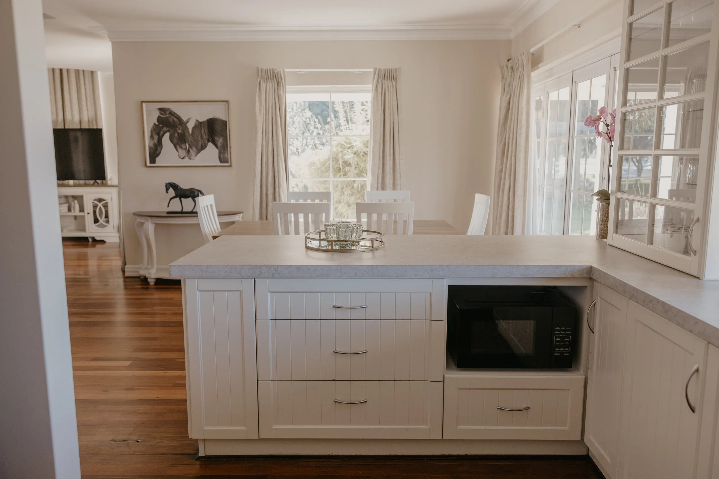 Interior view of a kitchen with white cabinets, a beige countertop, and a black microwave. The view extends to a dining area with a table and white chairs, large windows with curtains, and a decorative horse picture on the wall.