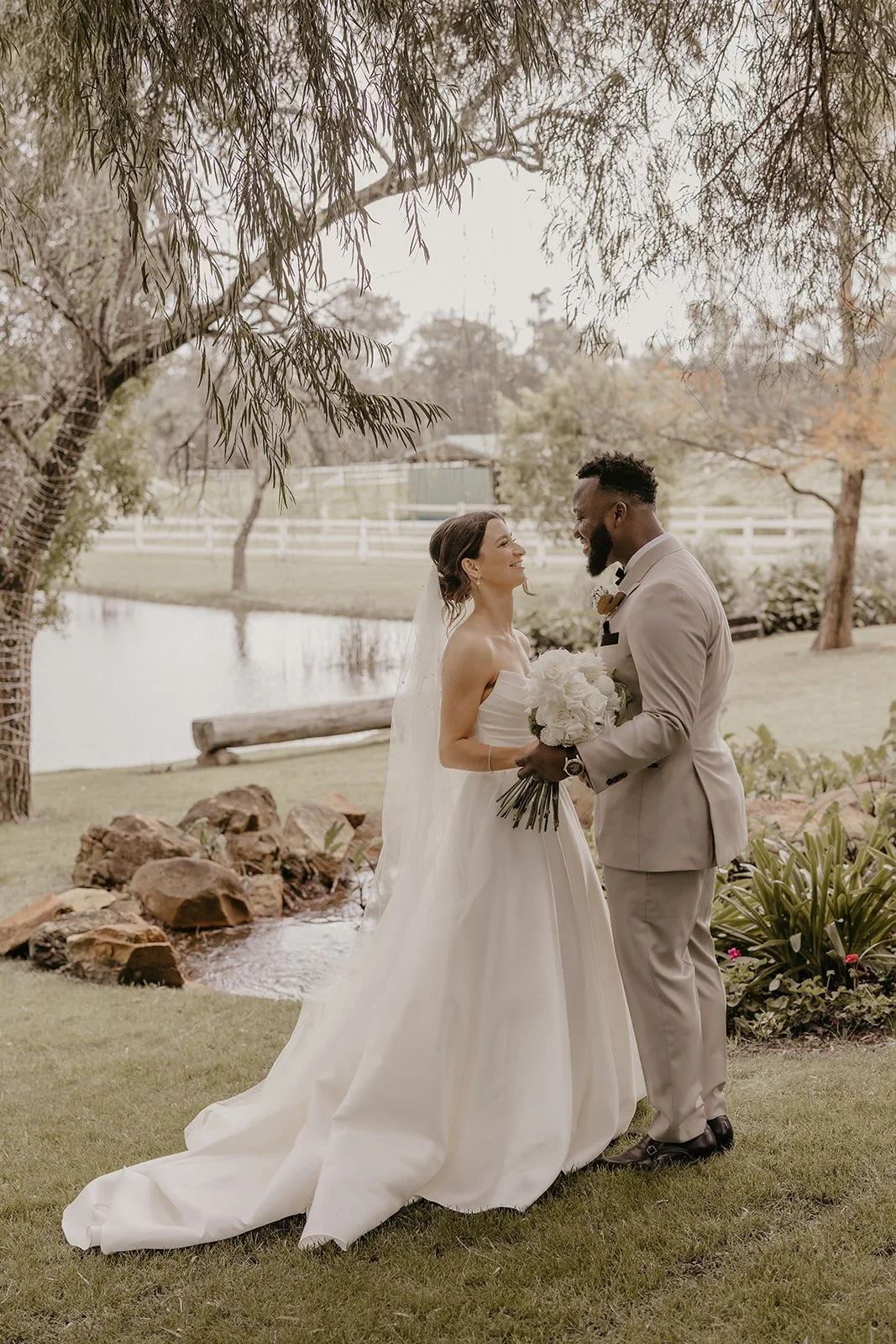 A bride and groom stand together outdoors near a pond, sharing a tender moment on their wedding day. The bride wears a strapless white wedding gown with a long train and veil, holding a bouquet of white roses. The groom wears a light grey suit, black