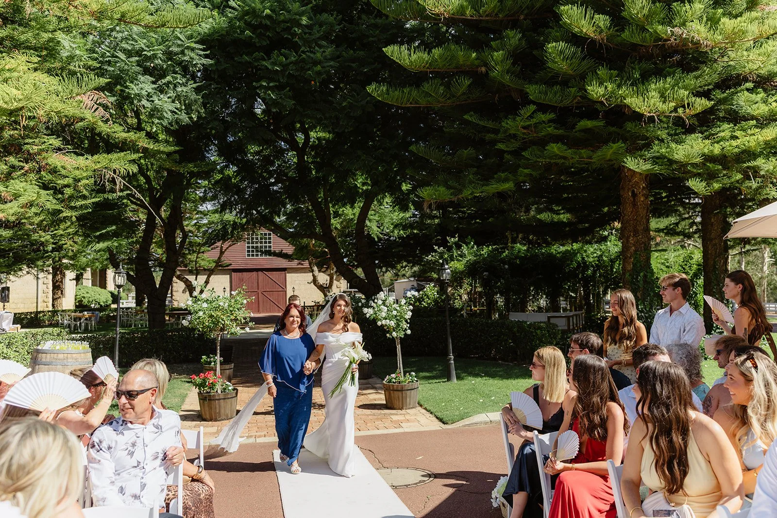 A wedding ceremony in a garden with a bride in a white dress walking on a white aisle, accompanied by a woman in blue. Guests are seated, many holding paper fans, and some are standing. The scene is shaded by large trees, with potted flowers and a ba