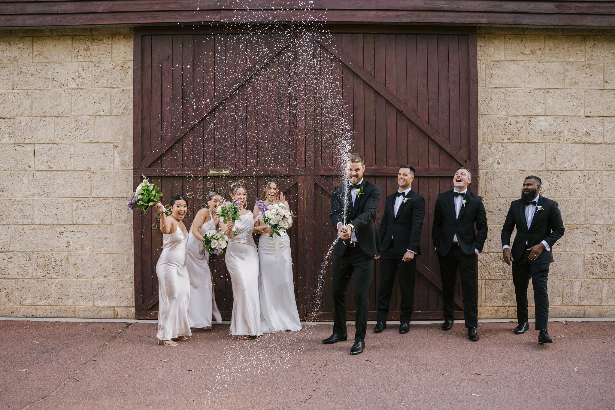 Group of wedding party members celebrating, with the groom in the center opening a champagne bottle, causing celebratory spray, while others cheer and laugh, standing in front of a large wooden barn door.
