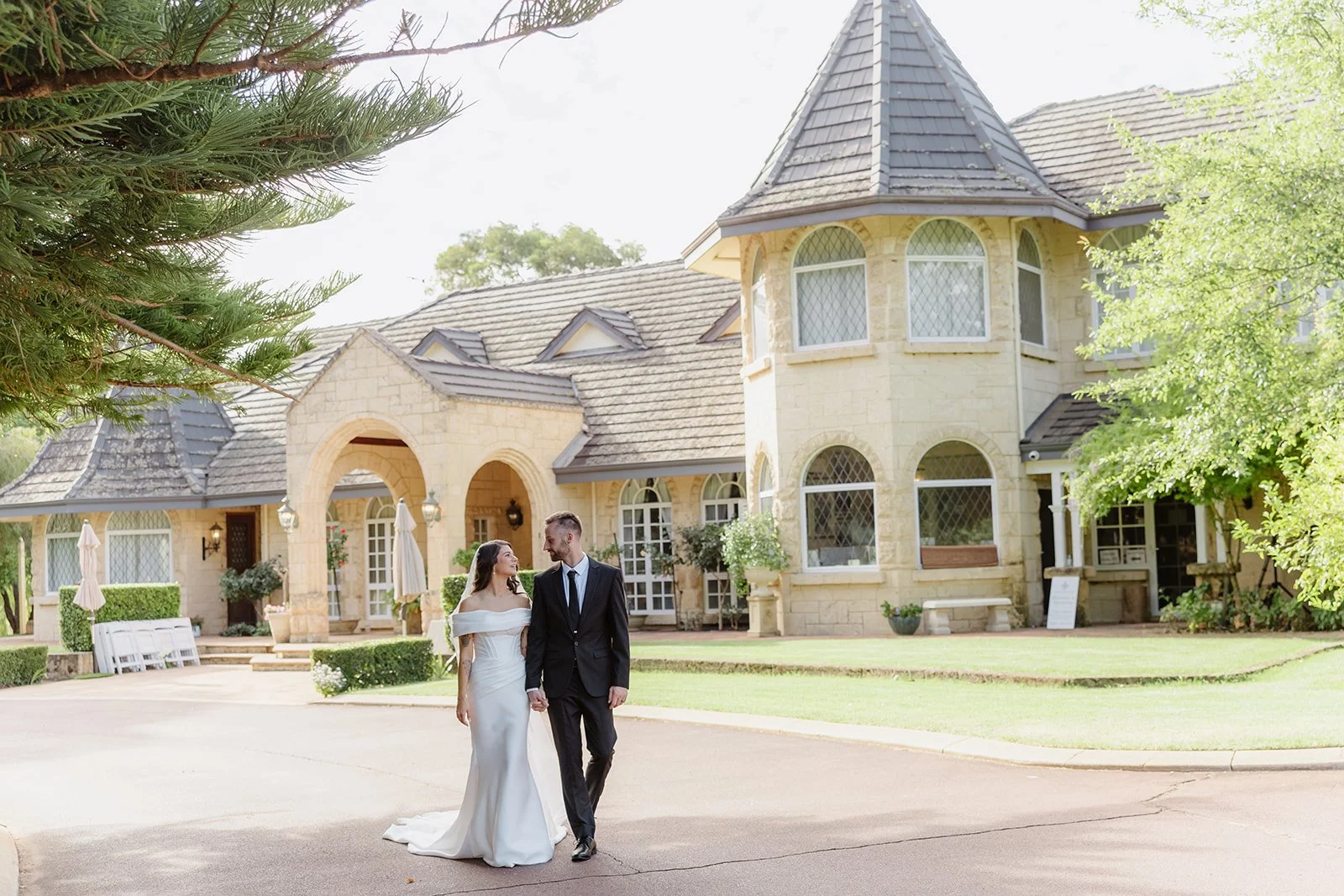 A bride and groom holding hands and walking outside a large, elegant stone mansion with a turret and arched windows, surrounded by green trees and well-manicured lawn.