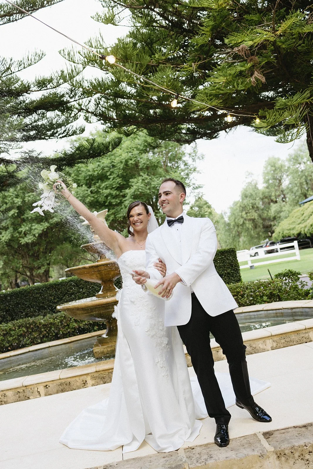 A newlywed couple celebrating outdoors by a fountain, with the bride tossing her bouquet and both smiling, surrounded by green trees and string lights.