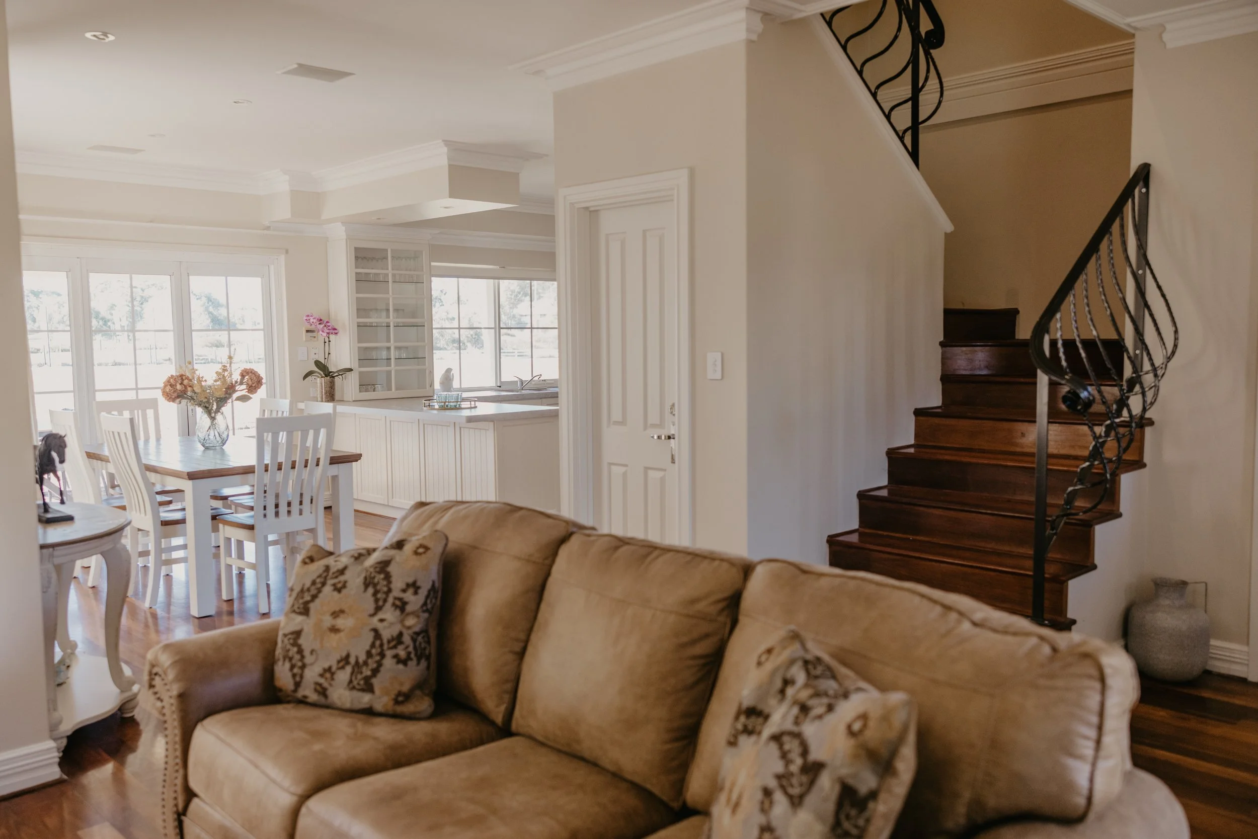 Living room area with a beige couch, decorated pillows, a dining table with chairs, floral arrangements, and a staircase with a decorative black wrought iron railing.