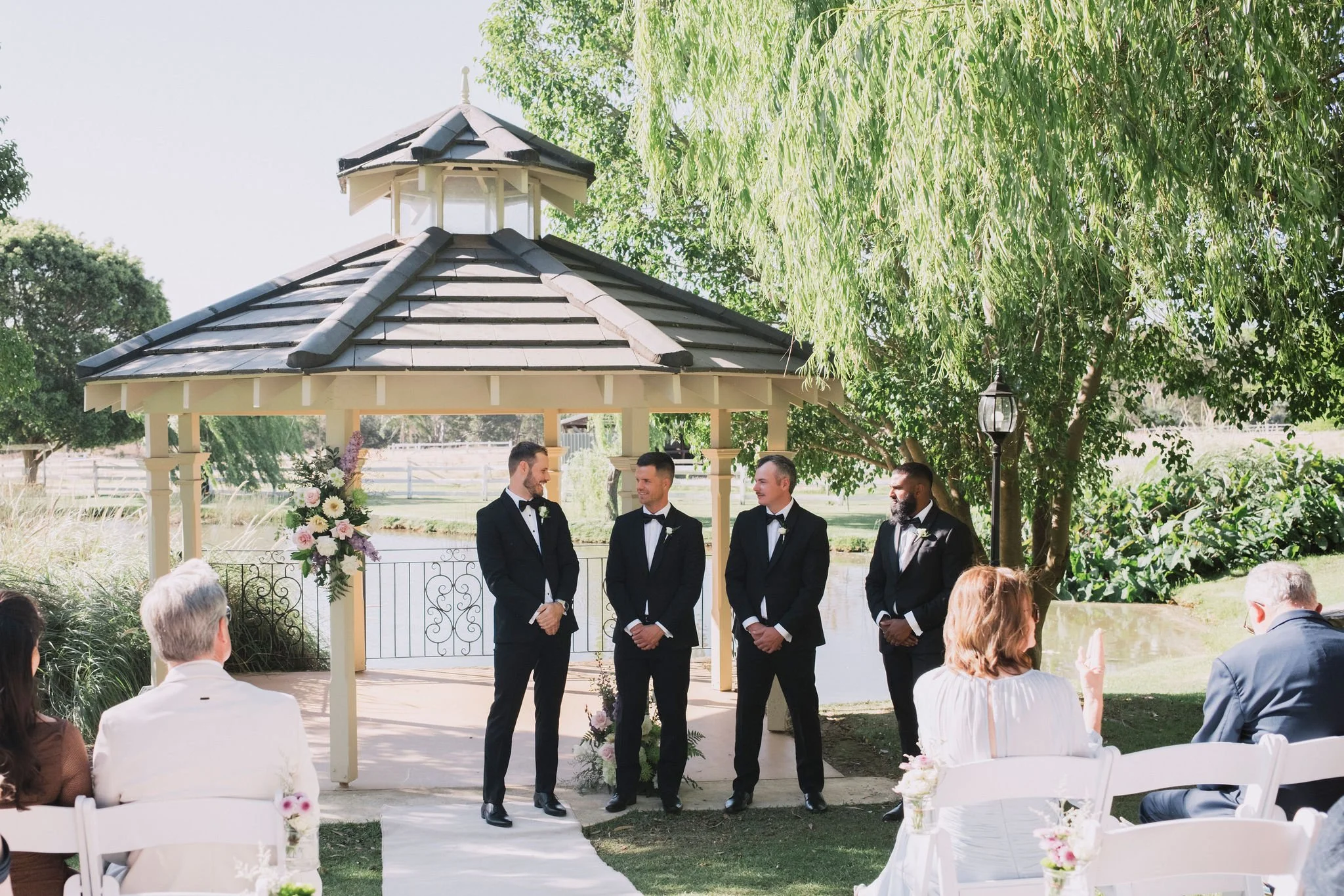 A same-sex wedding ceremony outdoors in front of a small pavilion by a pond, with four grooms in tuxedos, standing under a gazebo, and guests seated nearby.