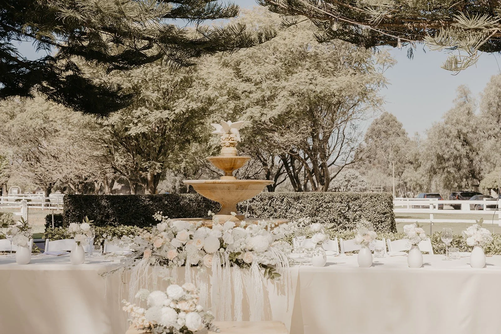 Outdoor wedding reception setup with a long table decorated with white floral arrangements and vases, in front of a fountain with trees and parked cars in the background.