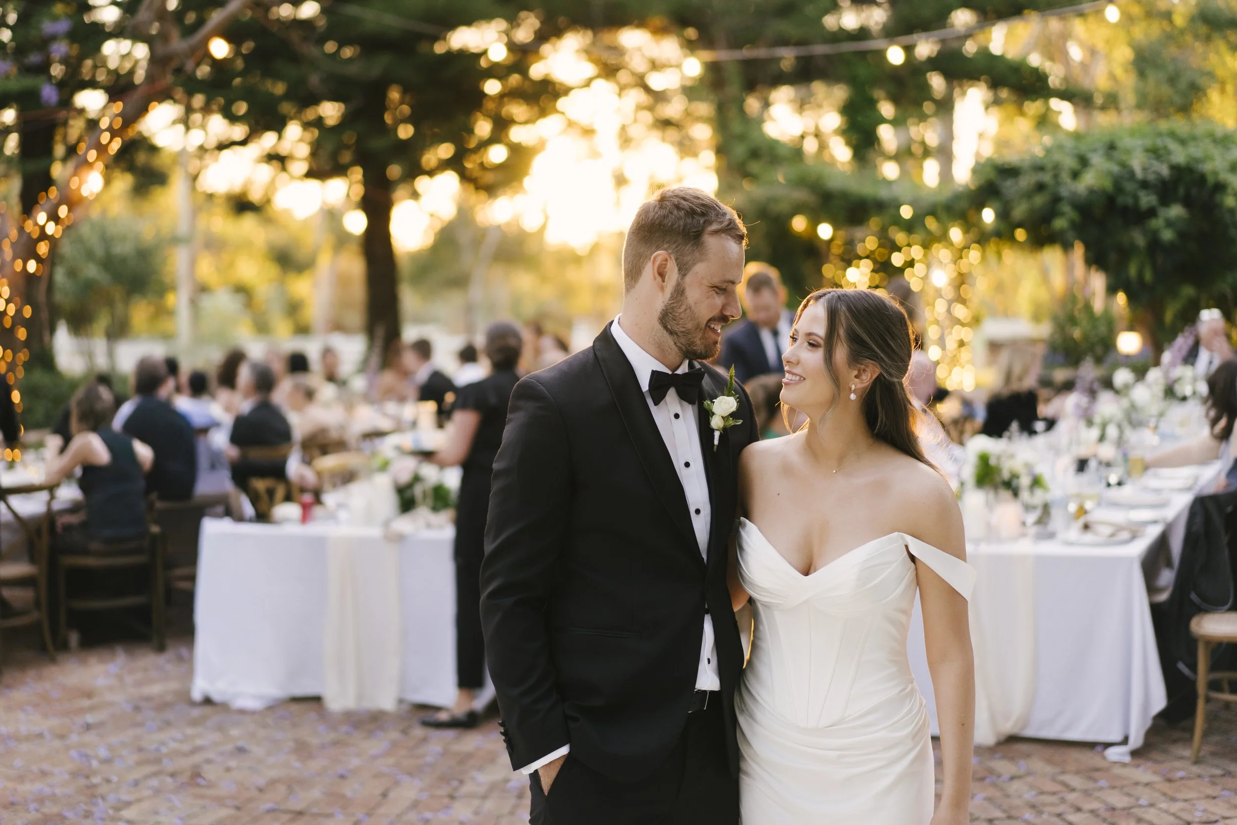 A happy couple, the bride in an off-shoulder wedding gown and the groom in a black tuxedo, sharing a moment at their outdoor wedding reception during sunset, with guests seated at decorated tables in the background.