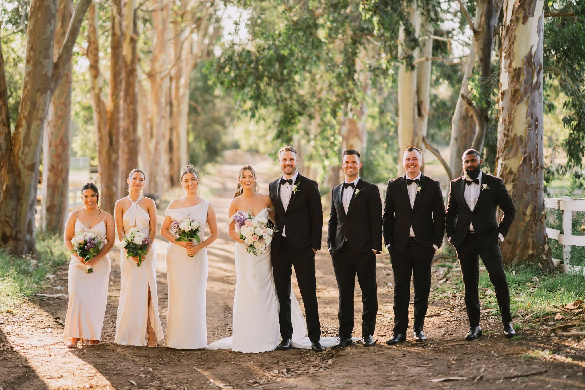 Wedding party walking on a dirt path lined with tall trees, including bridesmaids holding bouquets and groomsmen in tuxedos. The bride and groom stand in the center.
