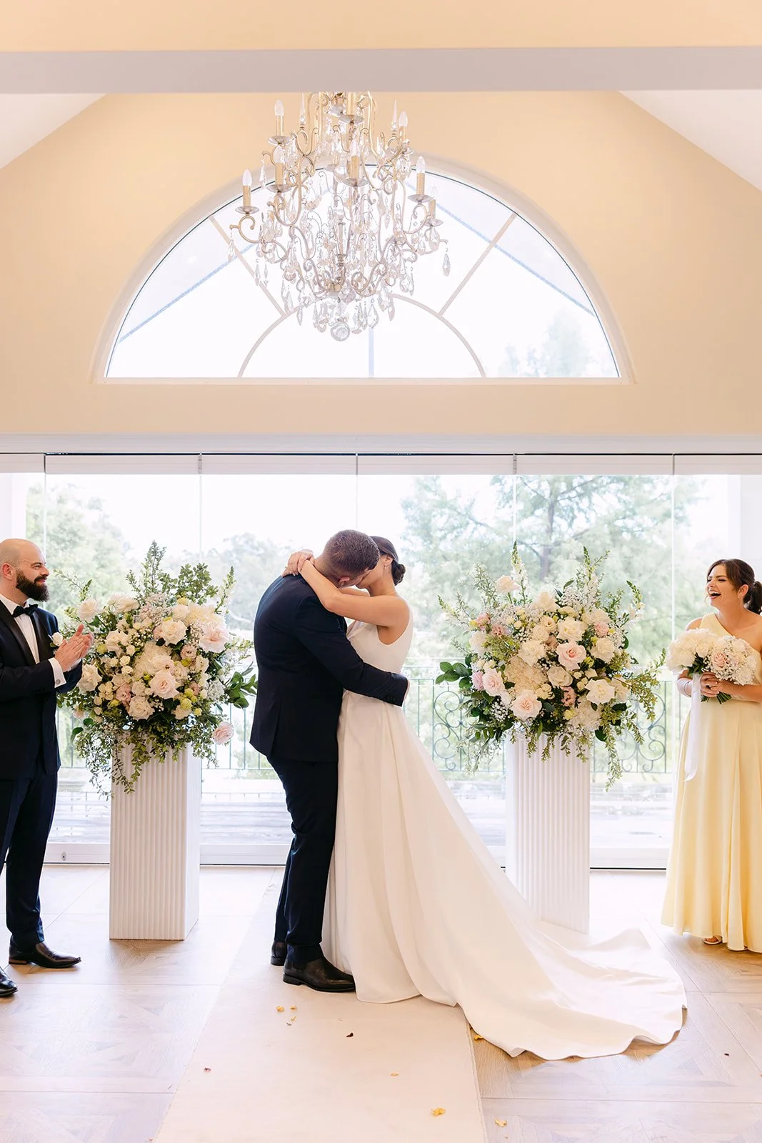 A bride and groom kiss during their wedding ceremony with friends clapping and smiling nearby, surrounded by large floral arrangements and a luxurious chandelier hanging above in a bright room with large windows.