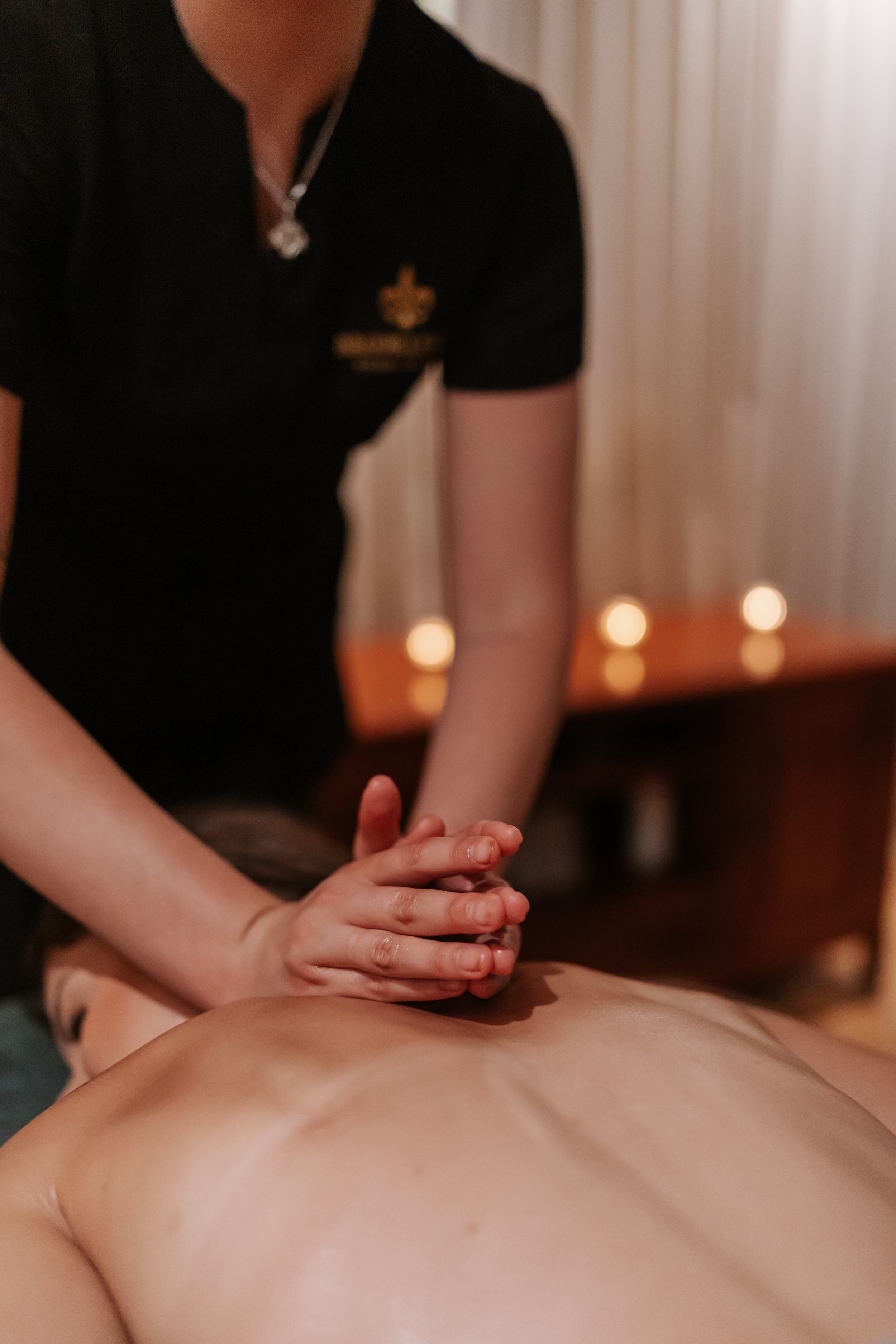 A person receiving a massage, lying face down on a massage table in a dimly lit room with candles, person giving the massage has a black shirt with a logo.