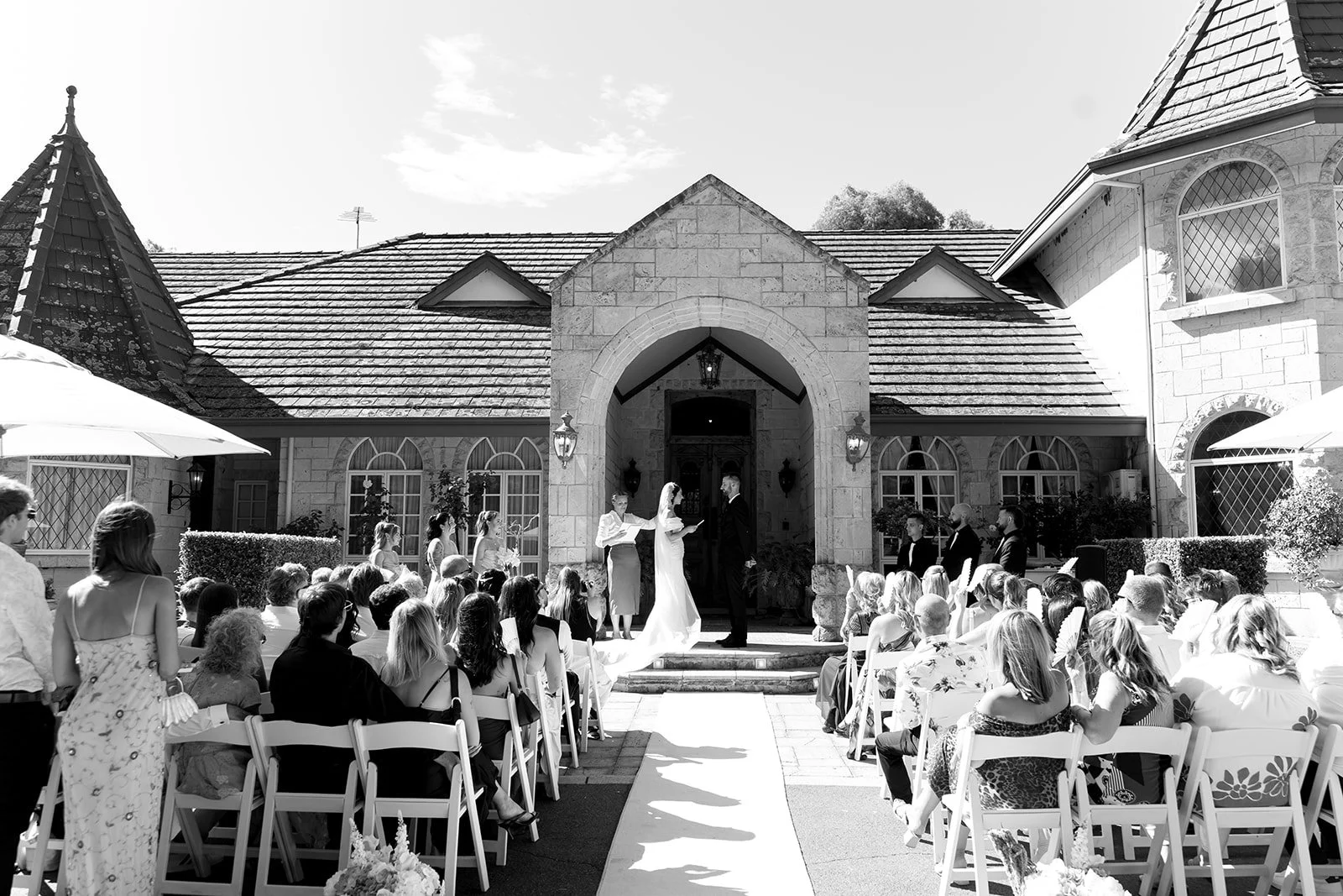 A wedding ceremony taking place outdoors in front of a large stone house with a porch. The bride and groom stand under an arch as the officiant and witnesses conduct the ceremony. Guests are seated in chairs on either side of an aisle, watching the e