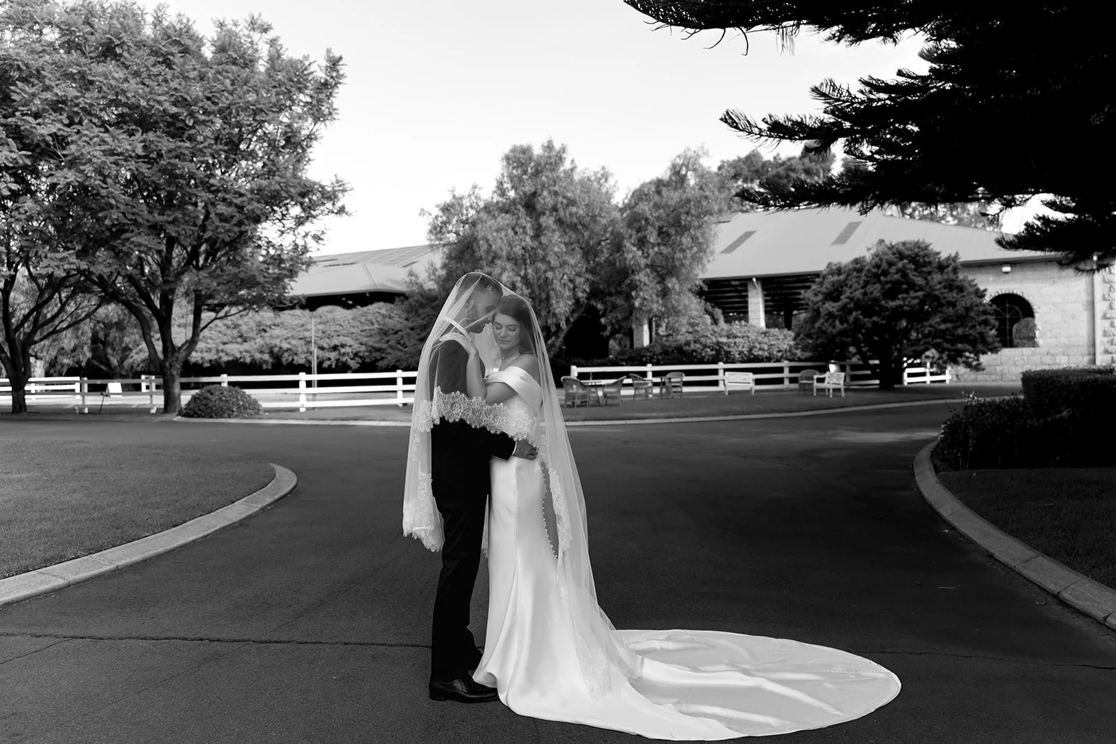 A bride and groom stand close together under a veil on a curved driveway, surrounded by trees and a building in the background, captured in black and white.