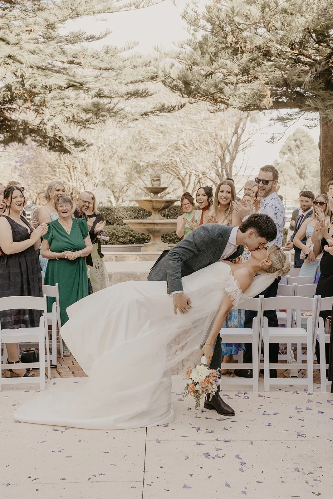 A newlywed couple sharing a kiss during their wedding ceremony while guests are clapping and smiling in the background, outdoors near a fountain and surrounded by trees.