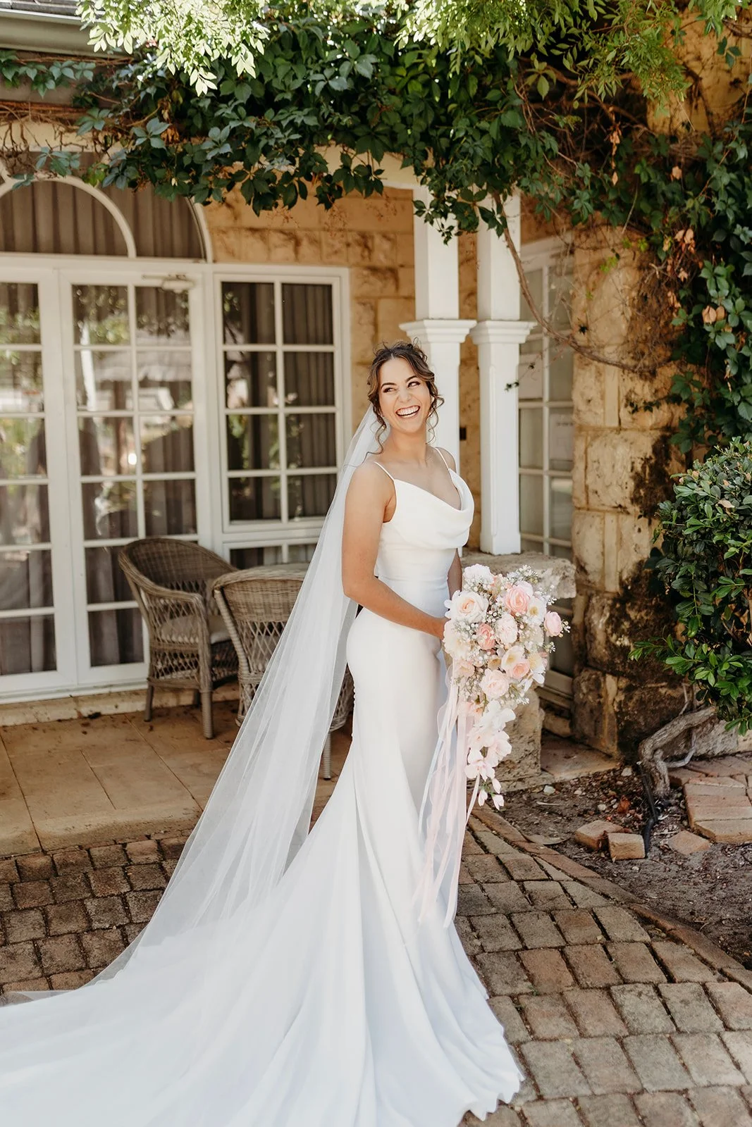 A bride in a white wedding dress, smiling and holding a pink and white flower bouquet, standing outside in front of a stone building with large windows and greenery.