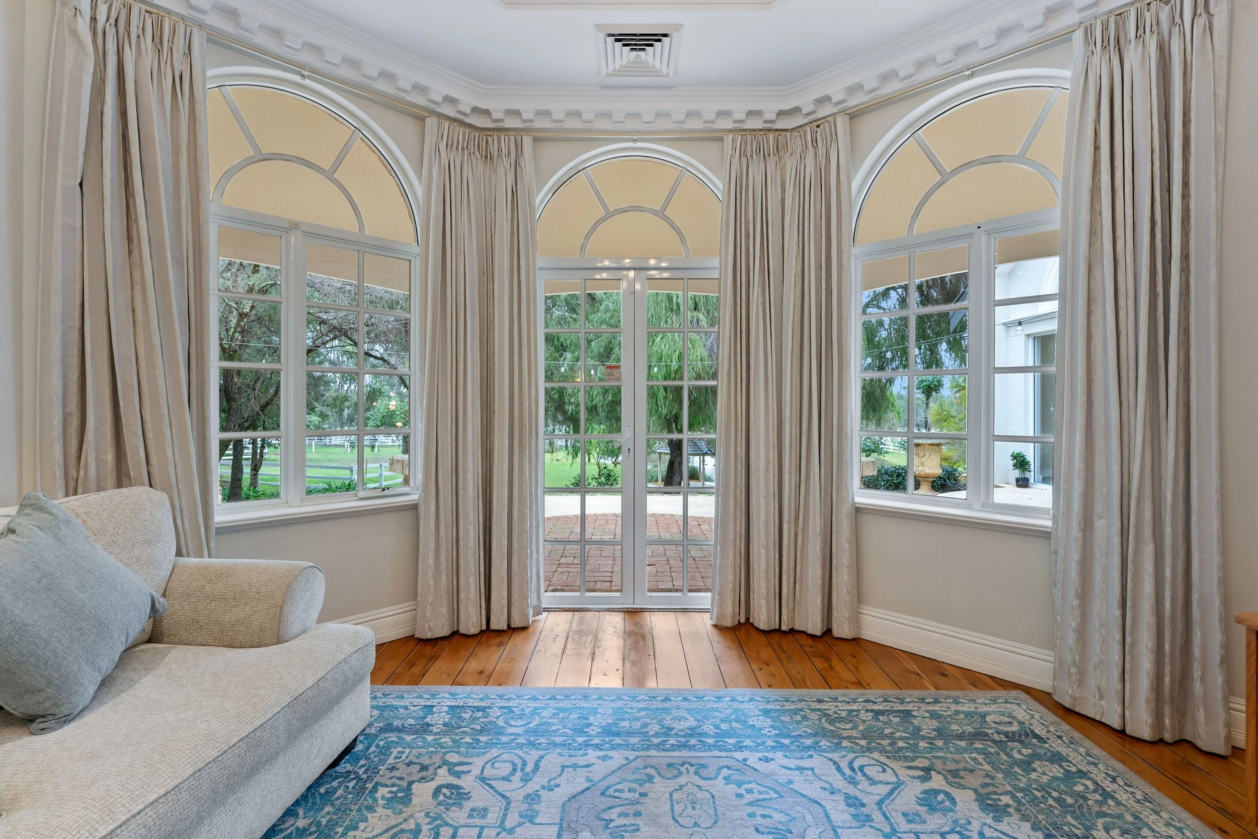 Interior view of a room with large arched windows and glass door, cream-colored curtains, hardwood floor, rug, and a cream sofa with blue cushions, overlooking a green outdoor area with trees.
