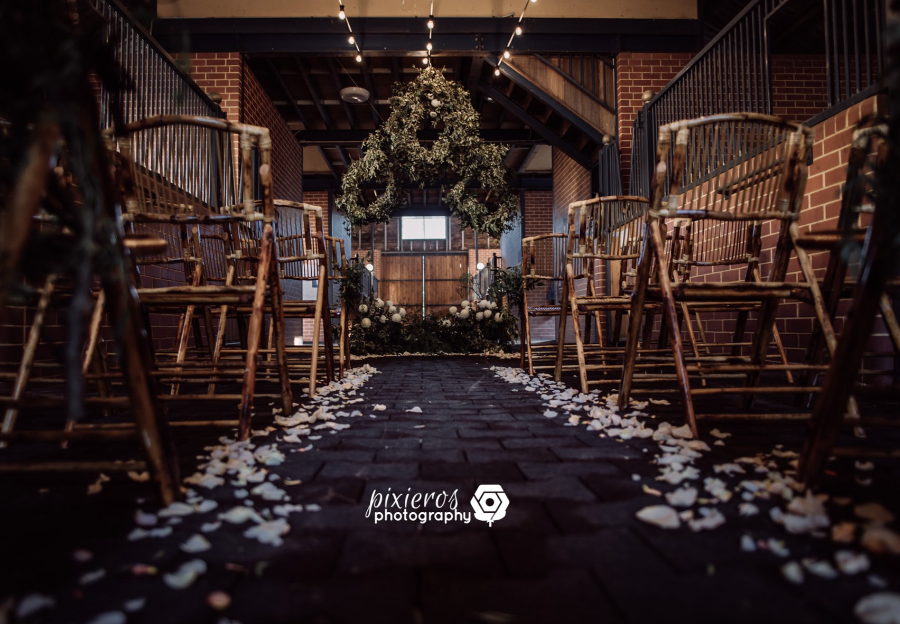 Indoor wedding ceremony setup with rows of wooden chairs arranged on both sides of a brick aisle, flower petals scattered on the floor, and a floral arch at the front, illuminated by string lights hanging from the ceiling.
