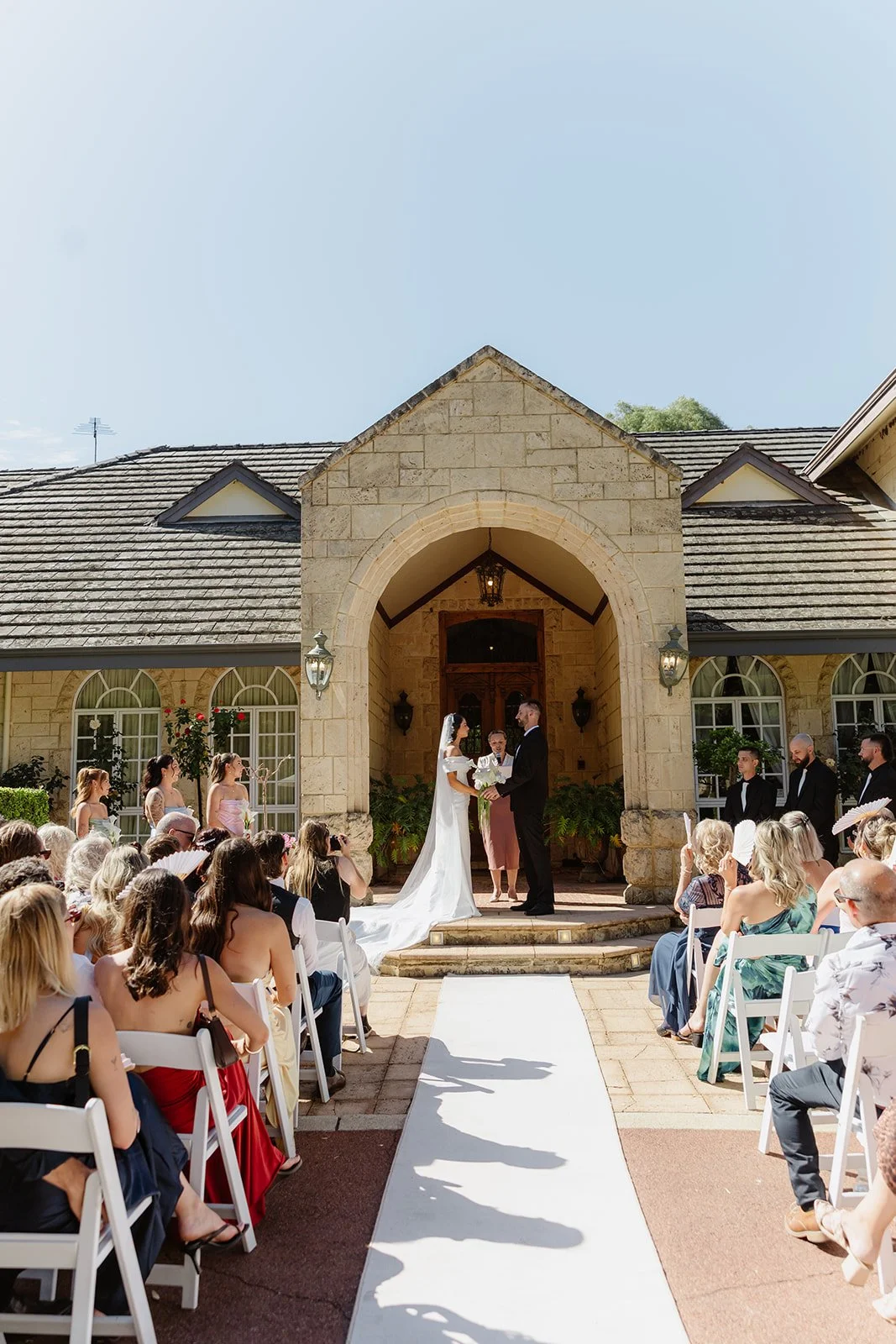 A wedding ceremony taking place outdoors in front of a stone building, with the bride and groom holding hands and exchanging vows, surrounded by guests seated on either side.