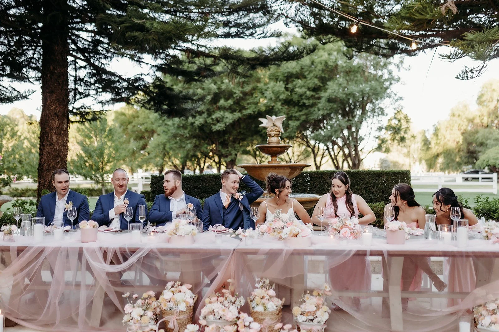 A wedding reception outdoor with eight people sitting at a decorated table in front of a fountain, surrounded by trees and greenery, during daytime.