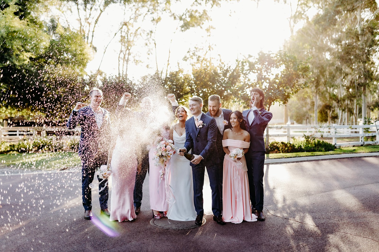 Group of people celebrating a wedding outdoors during sunset, with some holding bouquets, some laughing, and one person spraying champagne.