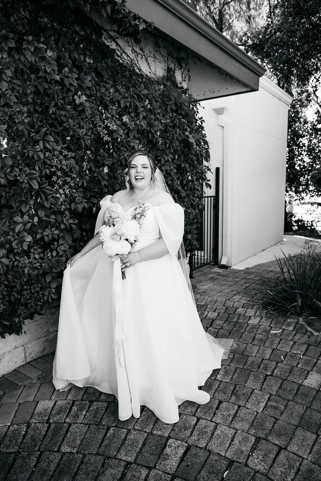 A woman in a wedding dress holding a bouquet, standing on a brick pathway next to a bush, making a playful face with one eye closed and tongue slightly out.
