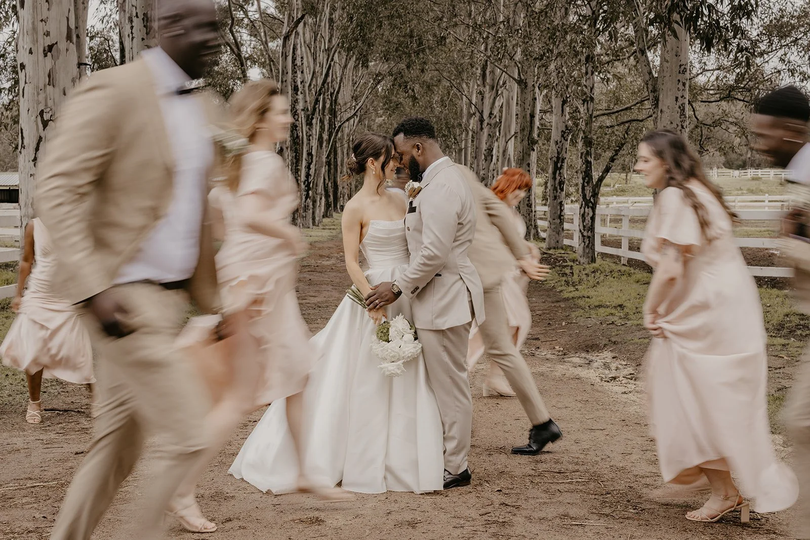 A wedding scene with a bride and groom standing close together, surrounded by bridesmaids and groomsmen walking past, in an outdoor setting with tall trees and a dirt path.