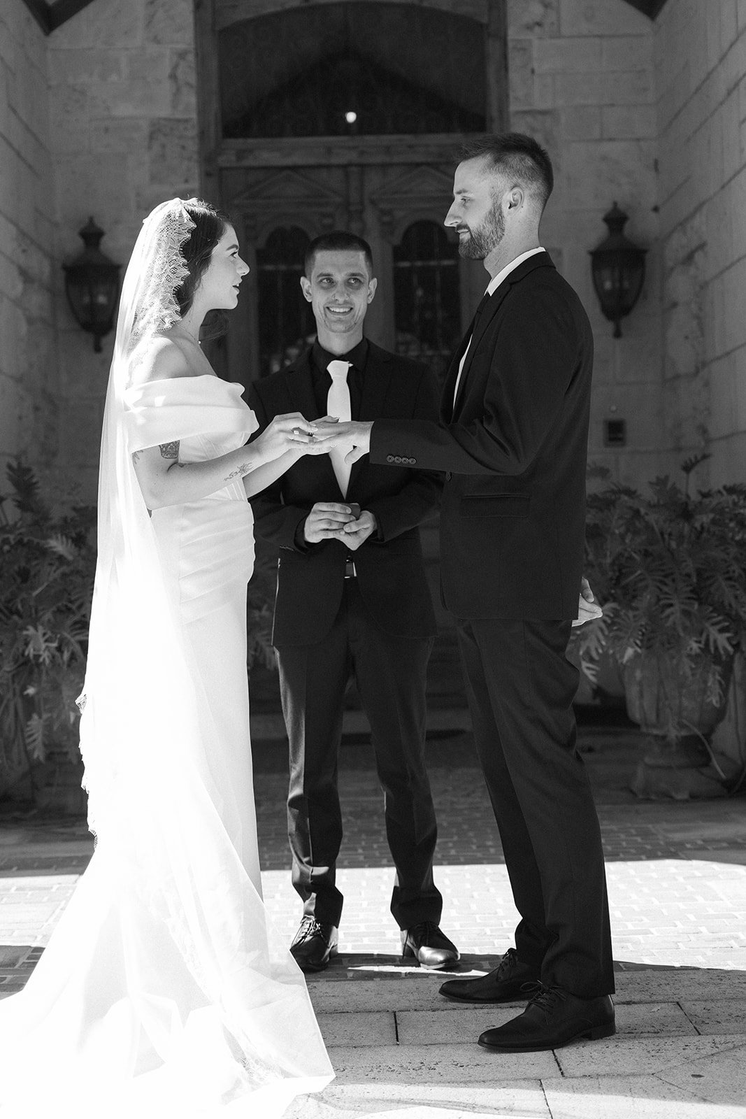 A black-and-white photo of a wedding ceremony with a bride and groom holding hands, standing face to face, with a minister in the background.