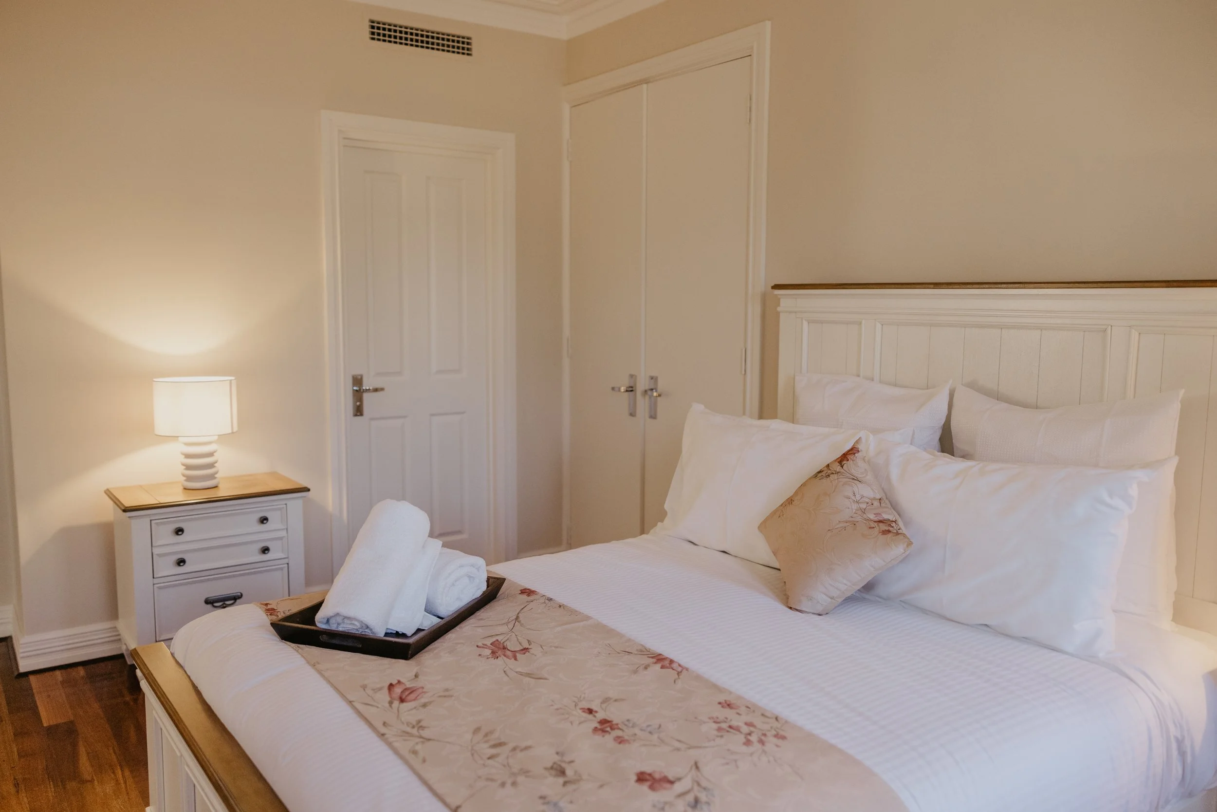 A cozy bedroom with a white bed, floral bed runner, and pillows. A tray with towels and toiletries is on the bed. There is a white nightstand with a lamp and a rolled towel. The room has beige walls, a white headboard, and a closet with double doors.