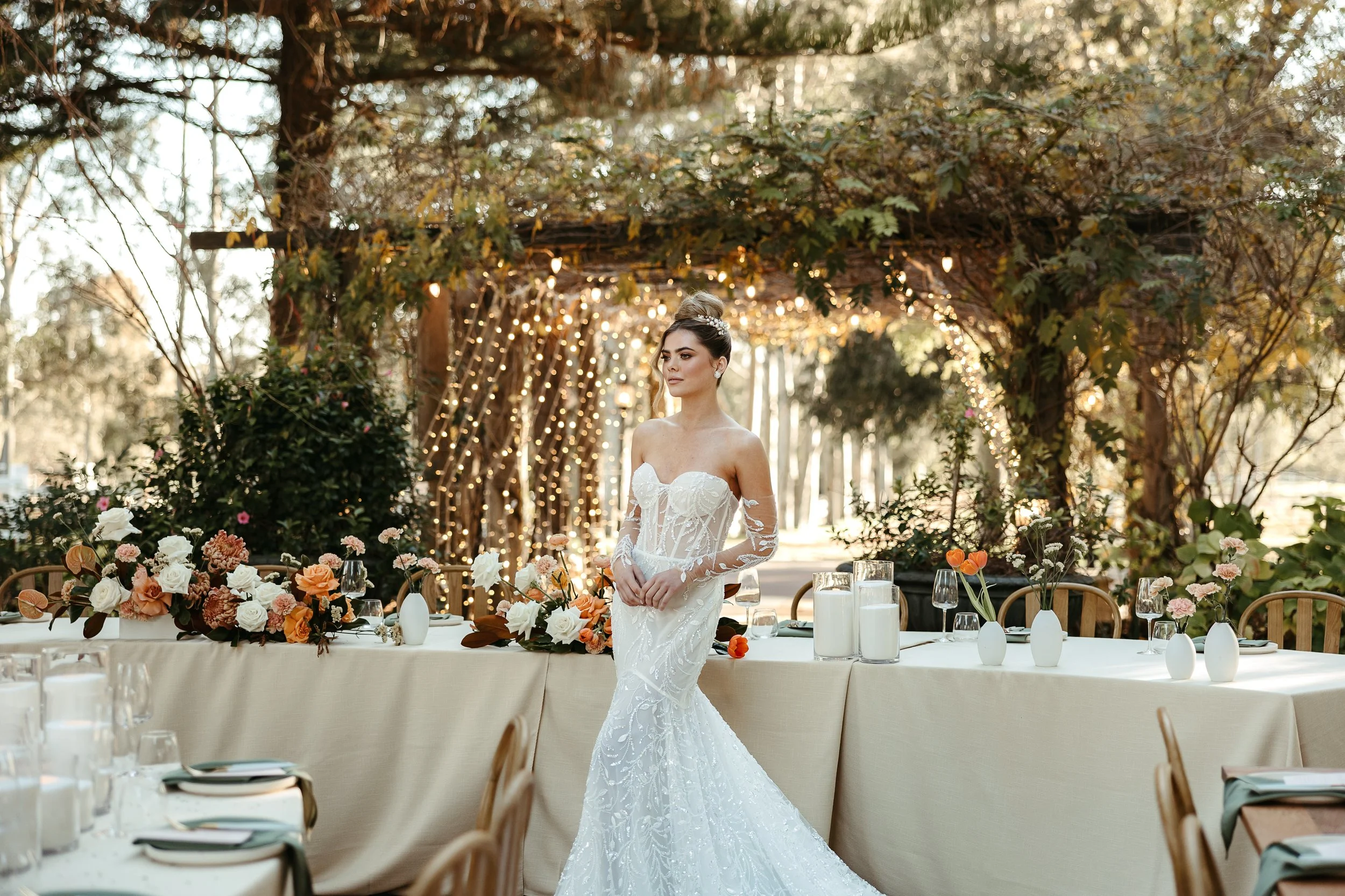 A bride standing in front of a decorated outdoor wedding reception area with fairy lights and a wooden archway, surrounded by flowers and tables set for guests.