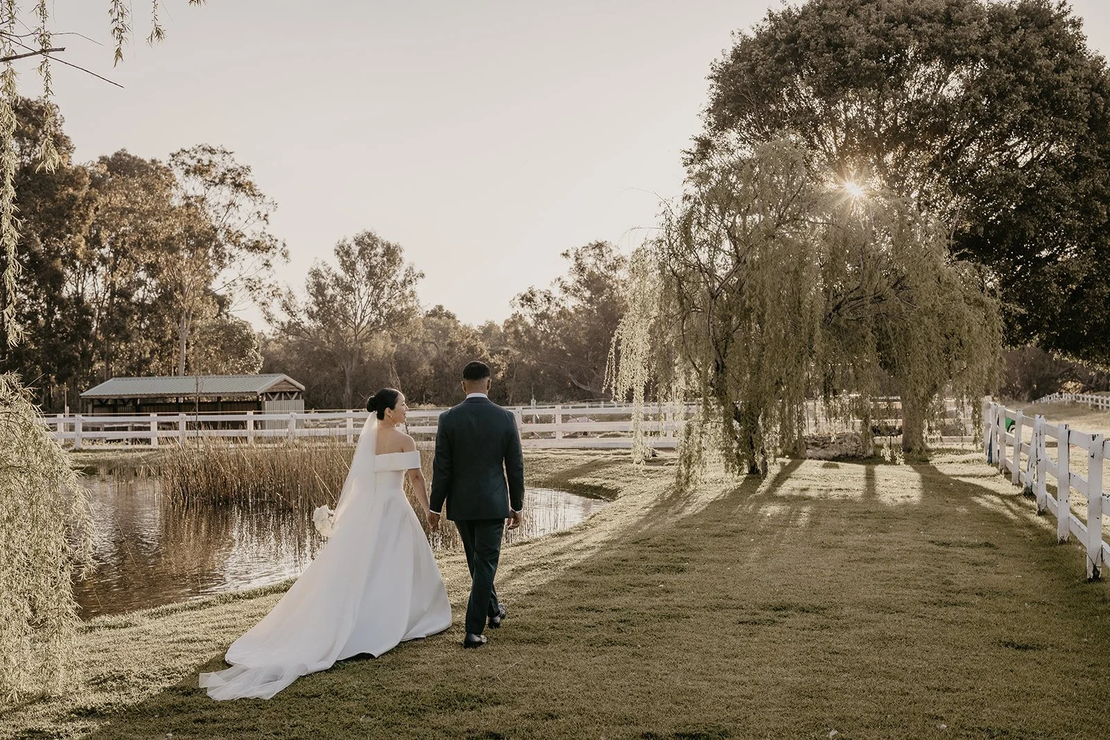 A bride and groom walking hand in hand near a pond with trees, sunlight shining through, and a wooden fence, on a serene outdoor wedding day.
