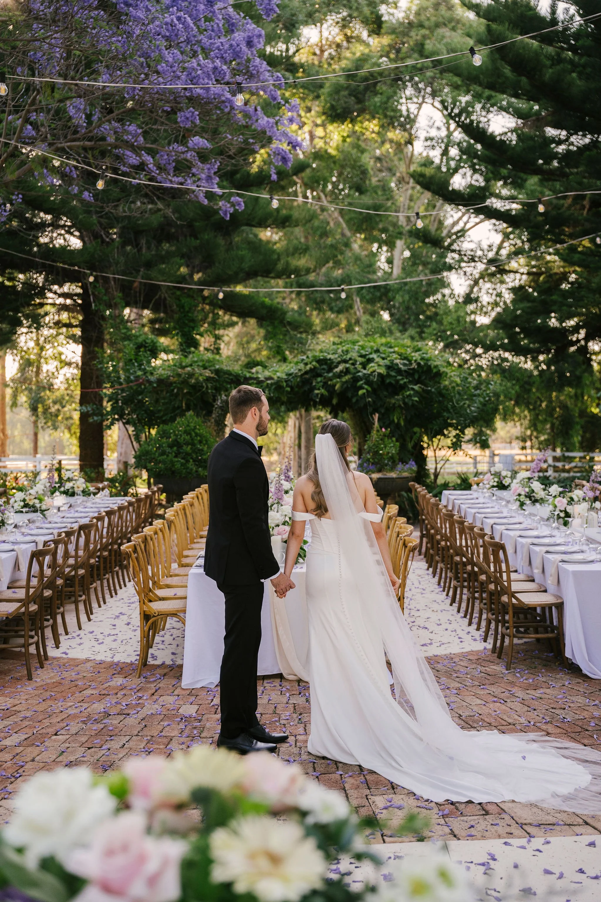 A bride and groom holding hands during their outdoor wedding ceremony in a garden, with flower-filled tables and string lights overhead.