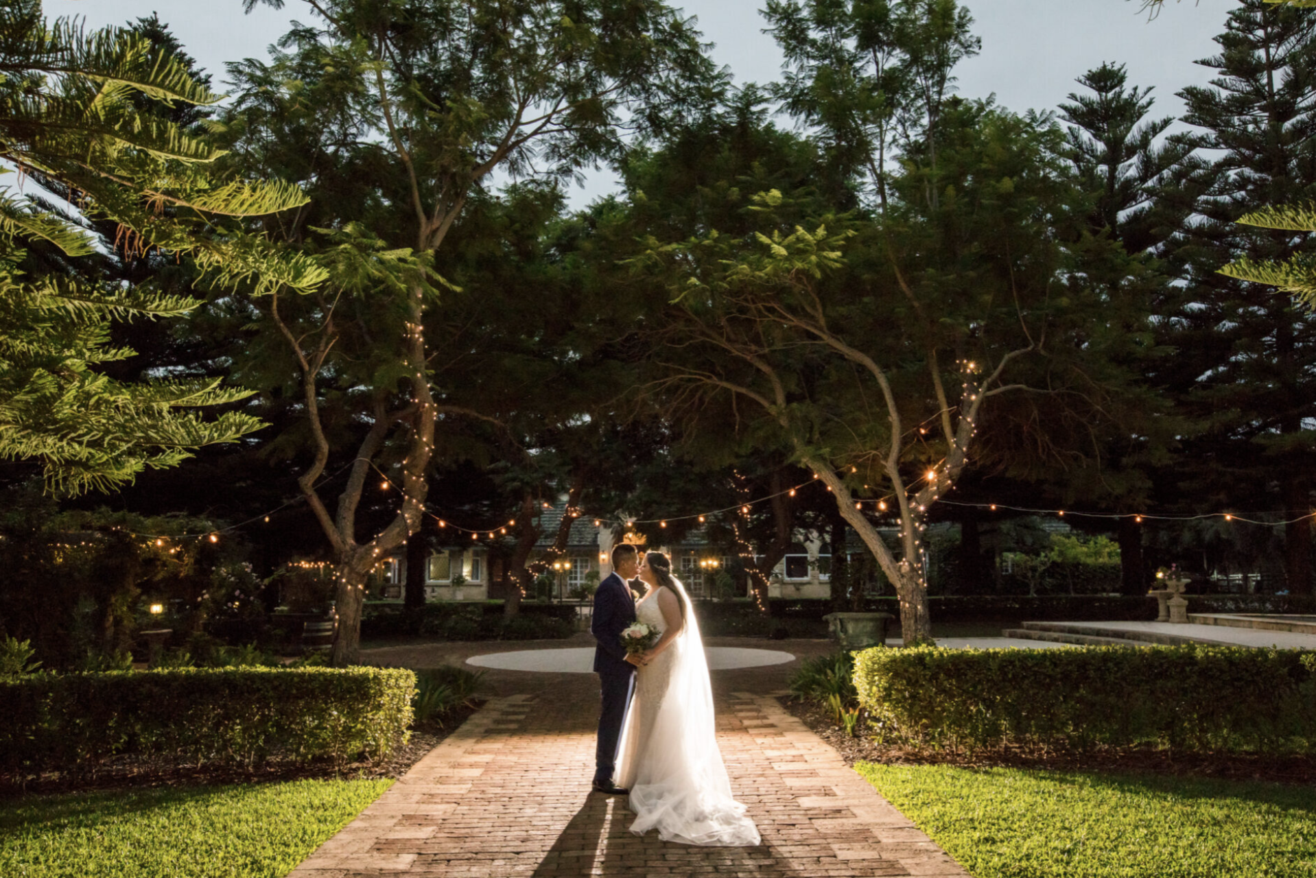 A couple dressed in wedding attire stand under a large tree decorated with string lights in a garden at dusk. The bride holds a bouquet, and the groom looks at her, with a backdrop of a house and surrounding greenery.