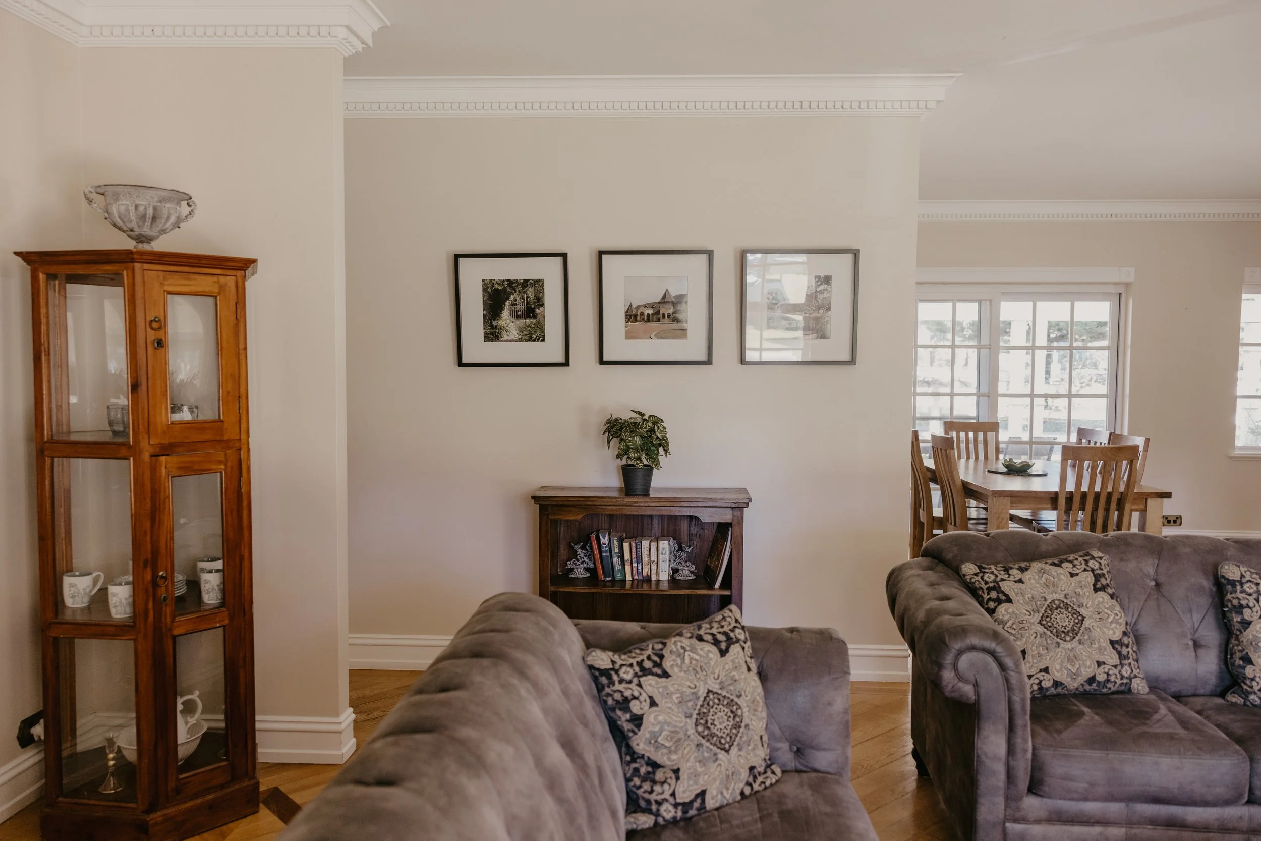 Living room with beige walls, crown molding, framed pictures, a wooden display cabinet, and furniture including a tufted sofa with patterned cushions and a wooden dining table with chairs near a large window.