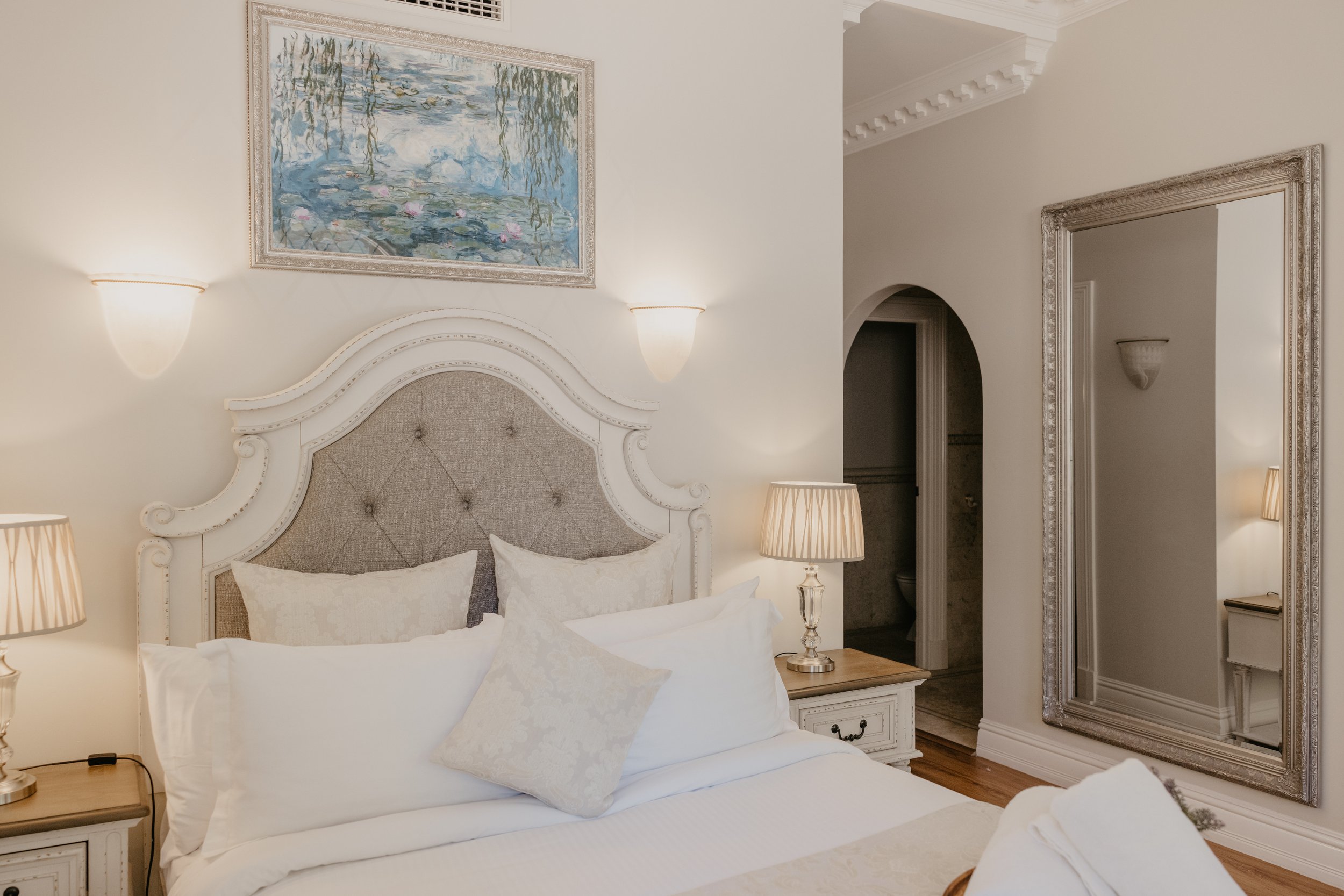 A bedroom with a white and beige upholstered headboard, white bedding, a beige throw pillow, and two bedside lamps with beige shades. There is a framed painting of water lilies hanging above the bed. To the right, there is a wall with a large mirror 