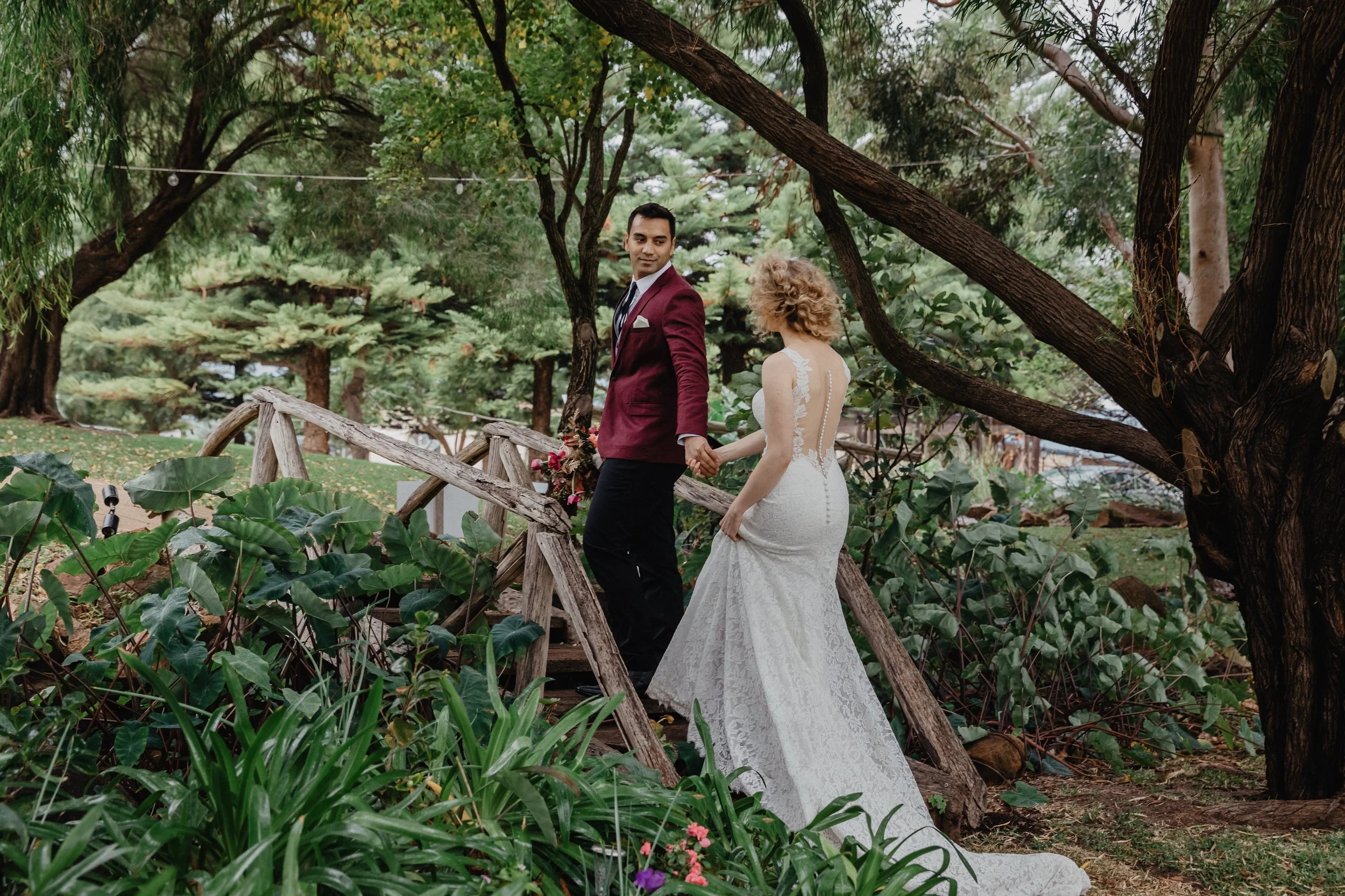 A bride and groom hold hands on a wooden bridge surrounded by lush greenery and trees.