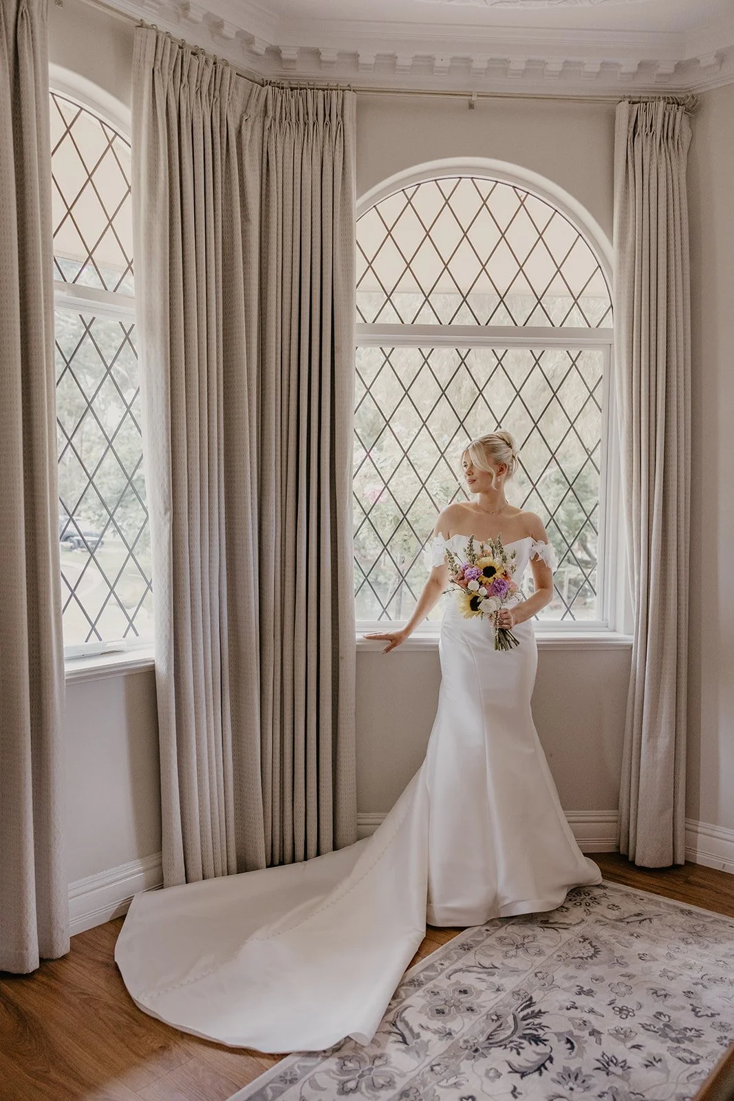 A bride in a white wedding gown holding a bouquet of flowers, standing by a large window with diamond-shaped panes and surrounded by beige curtains in a well-lit room.