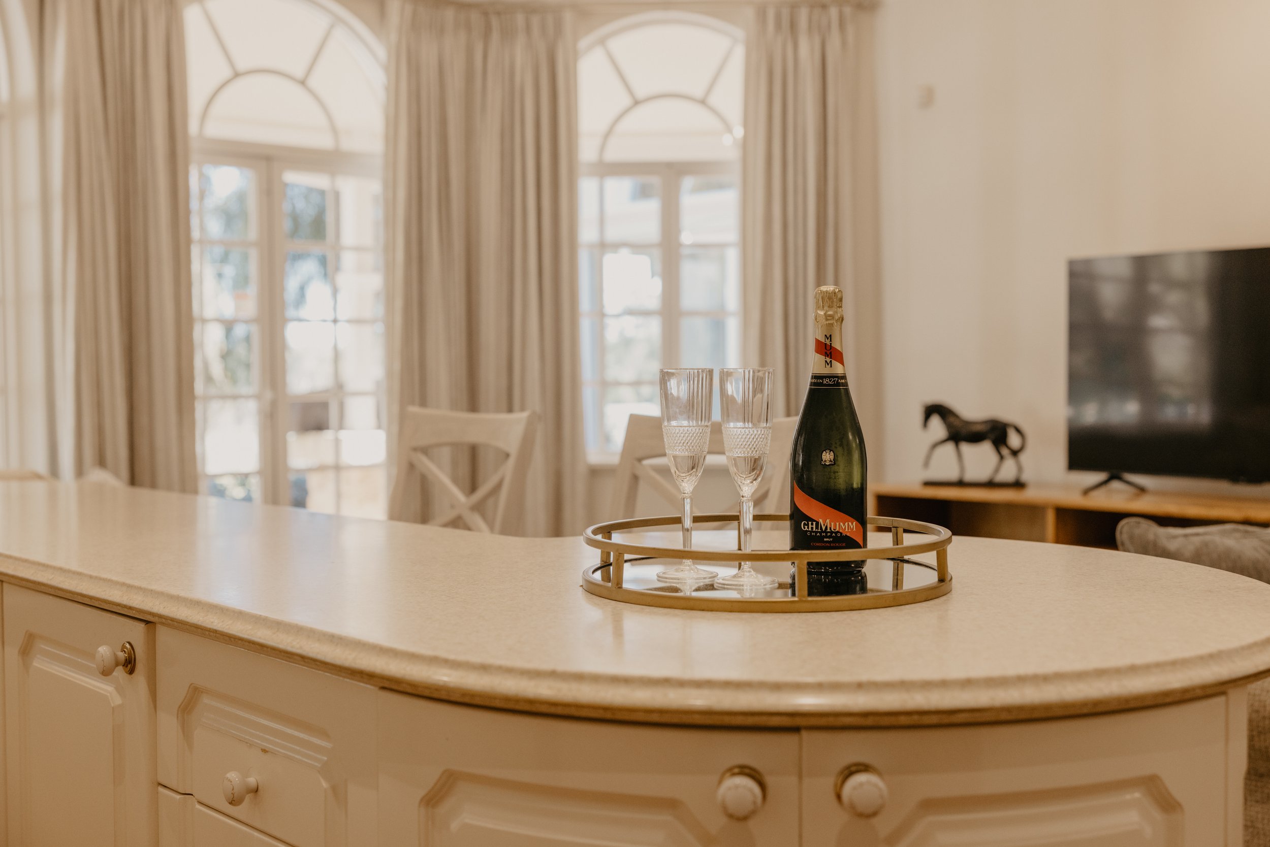 A kitchen island with a tray holding a bottle of champagne and two champagne glasses, with a background of large windows with curtains and a living room with a television and decorative horse figurine.