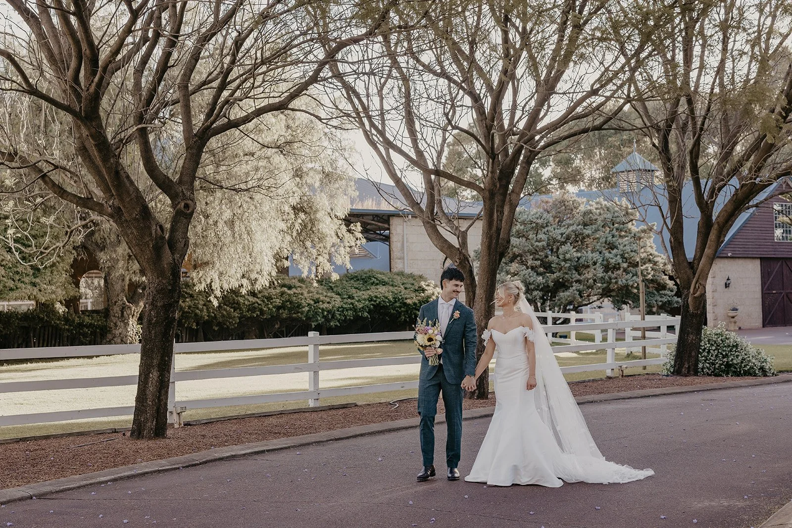 A bride and groom walking hand in hand on a paved road, with trees and a white fence in the background, during their wedding day.