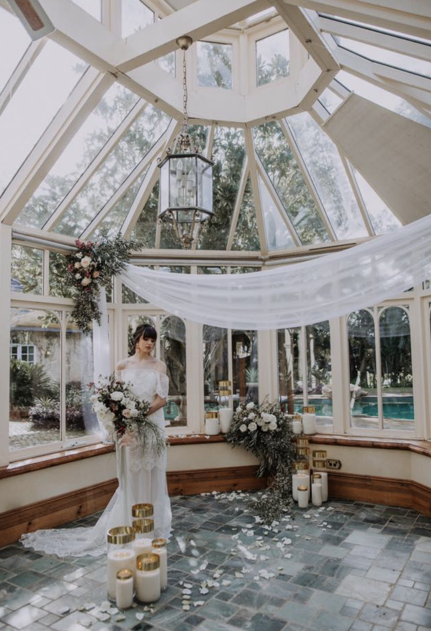 A bride in a white wedding dress holding a large bouquet stands inside a glass conservatory decorated with white flowers, candles, and white drapes for a wedding ceremony.