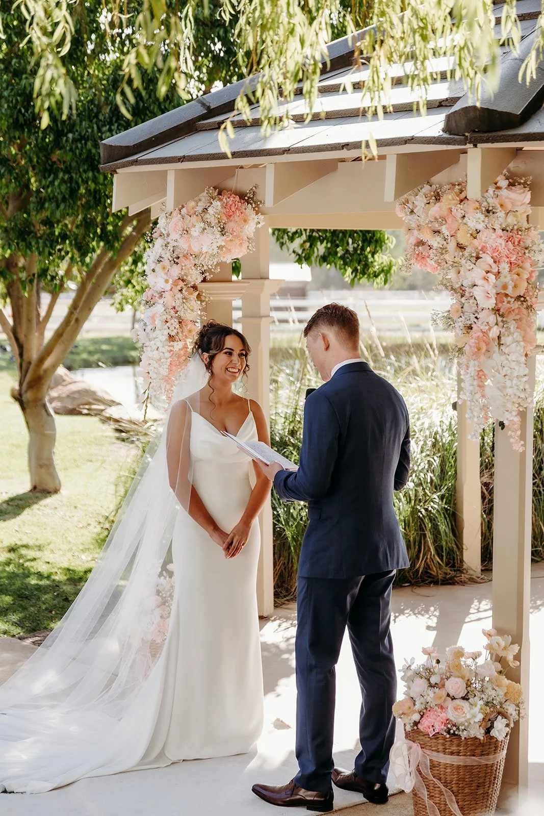 A bride and groom exchanging vows under a floral decorated arch outdoors on a sunny day.