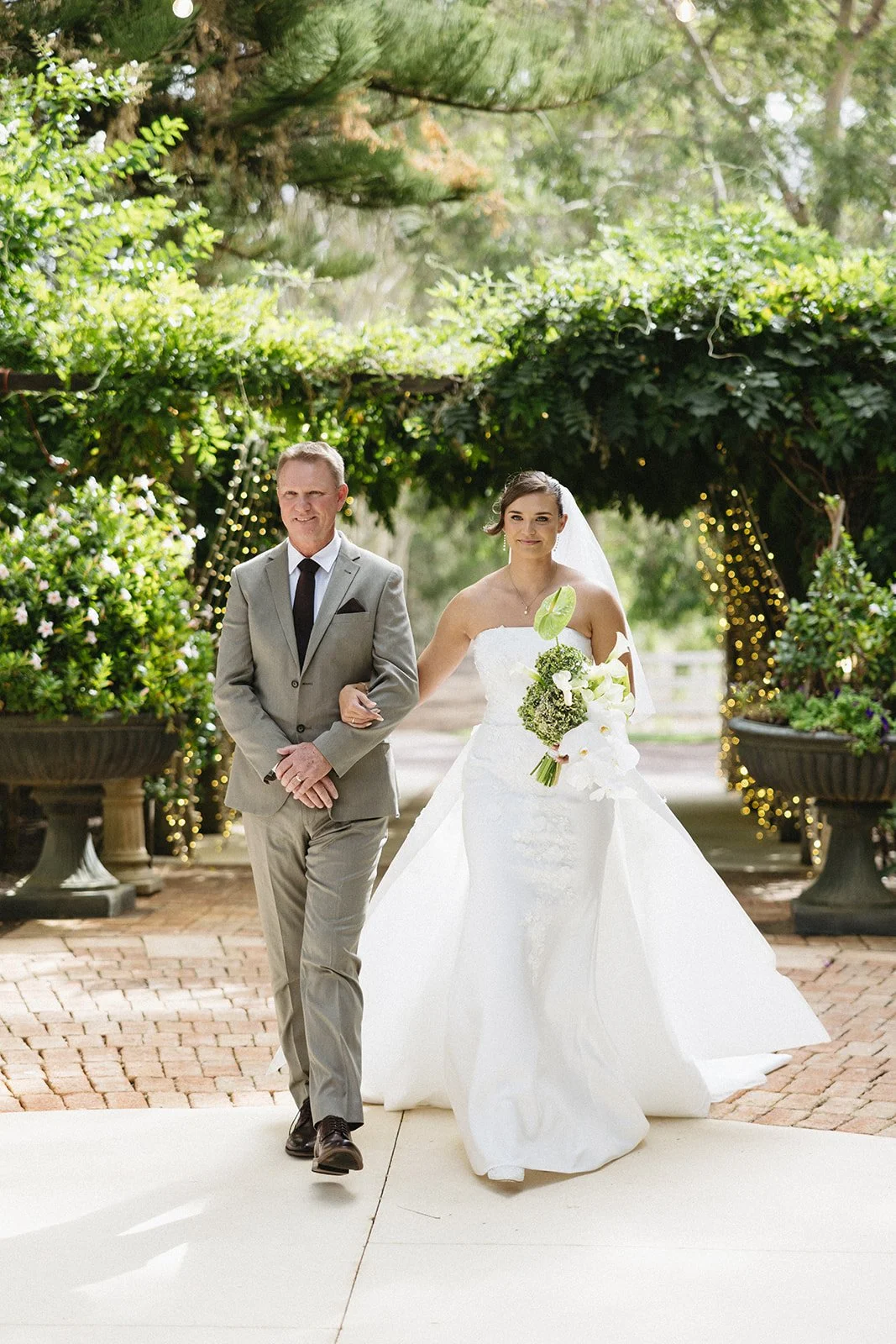 A bride in a white wedding gown holding a bouquet, walking arm-in-arm with a man in a gray suit, outdoors in a garden setting decorated with string lights and large flower pots.