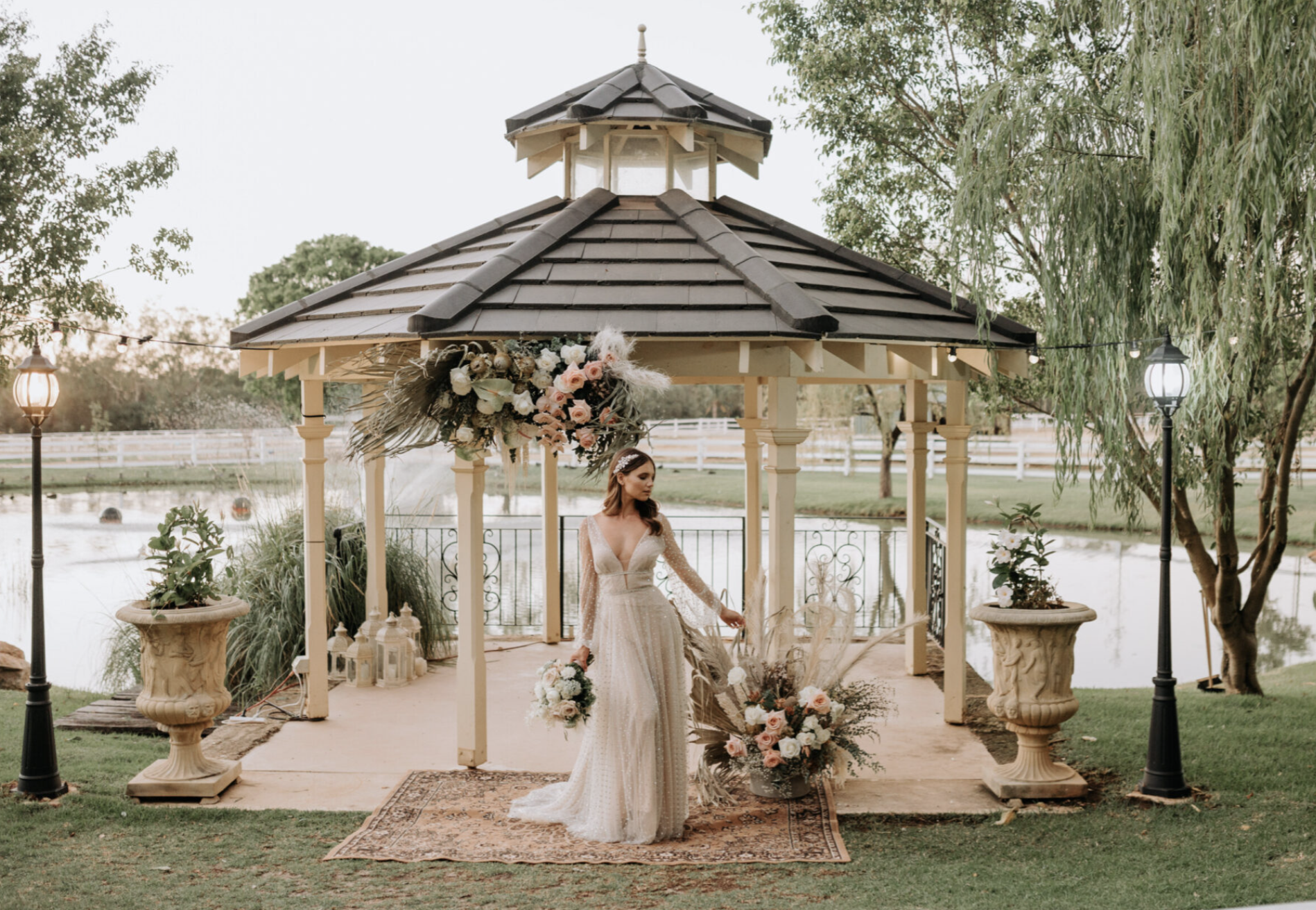 Bride in a white wedding dress standing on a rug under a gazebo decorated with floral arrangements near a pond with a fountain, surrounded by trees and outdoor lighting.