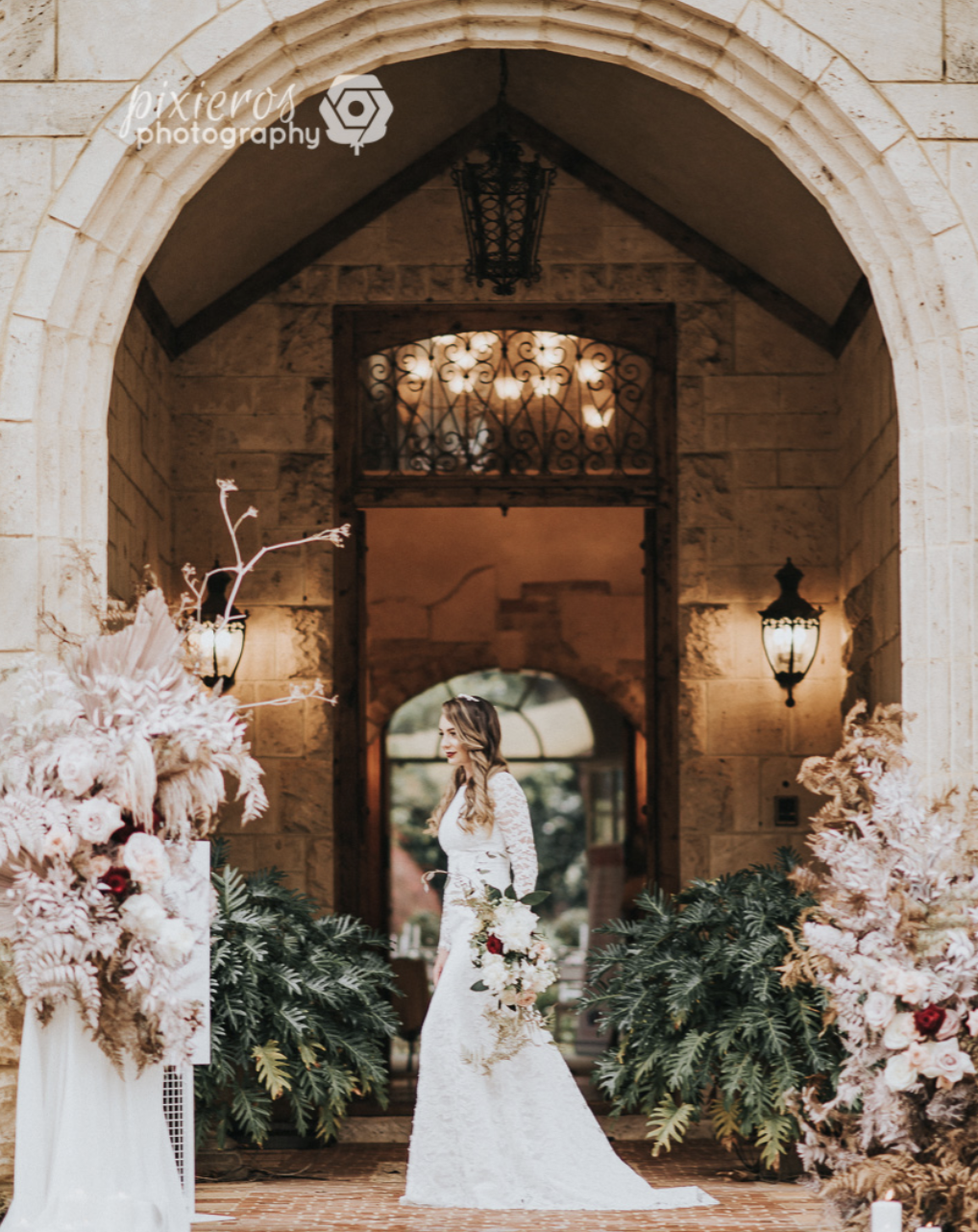 A bride in a white lace wedding dress holding a bouquet of flowers, standing in front of a stone archway with floral arrangements on either side.