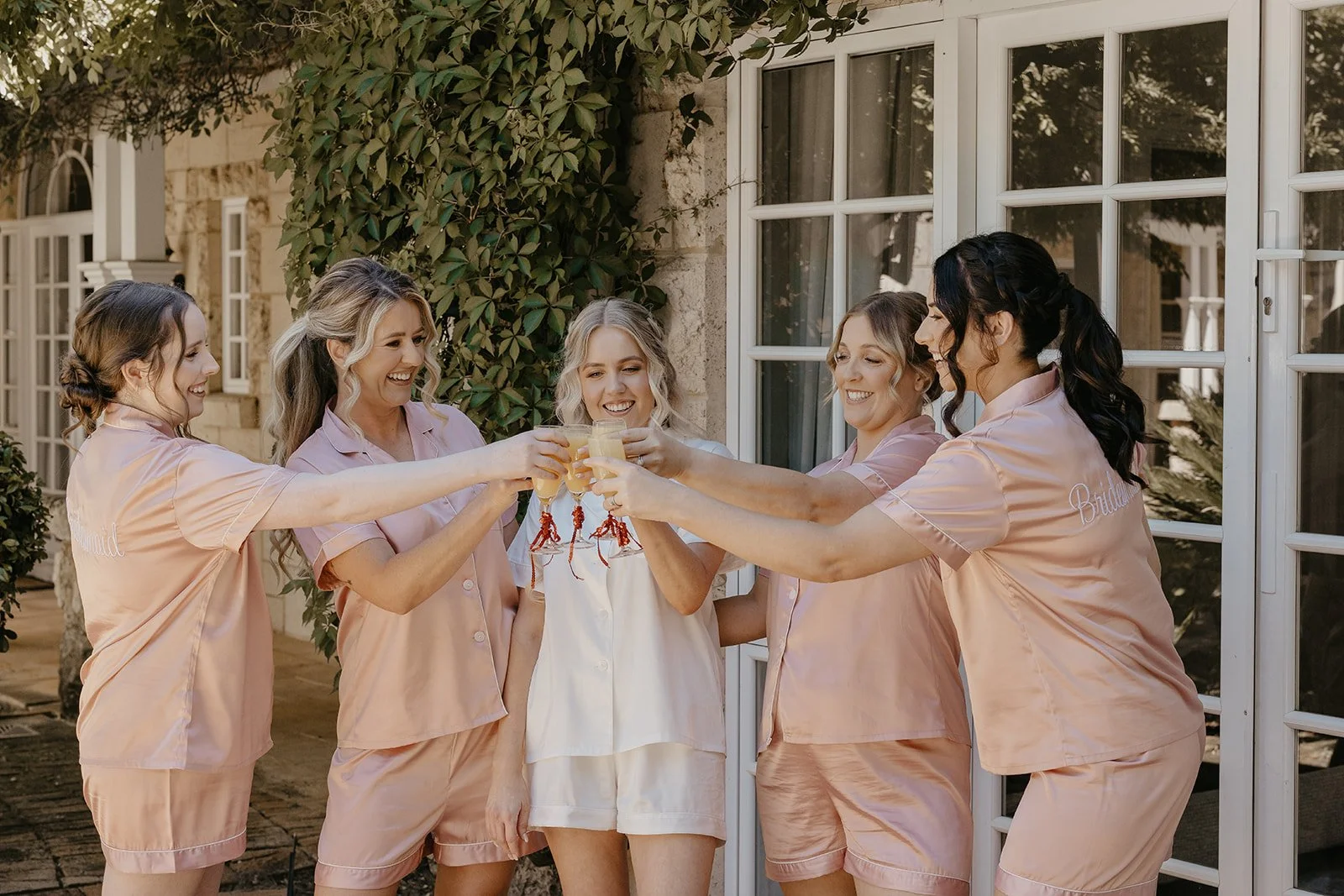 A group of six women in matching pink pajamas celebrating with glasses of champagne outside a house.