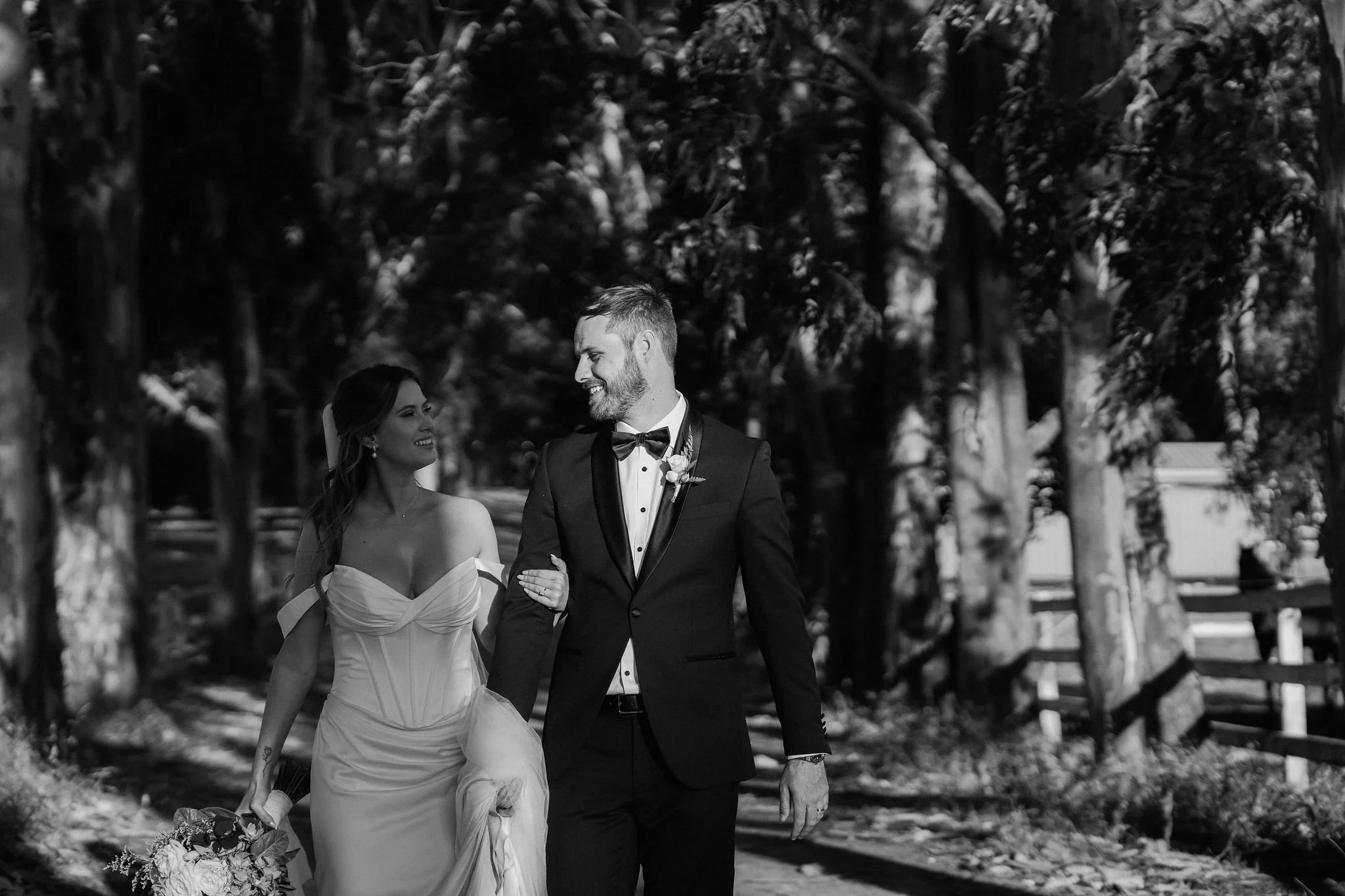 A black-and-white photograph of a newlywed couple walking arm in arm in a wooded area. The bride is wearing a strapless wedding gown and holding a bouquet, while the groom is in a tuxedo with a bow tie.